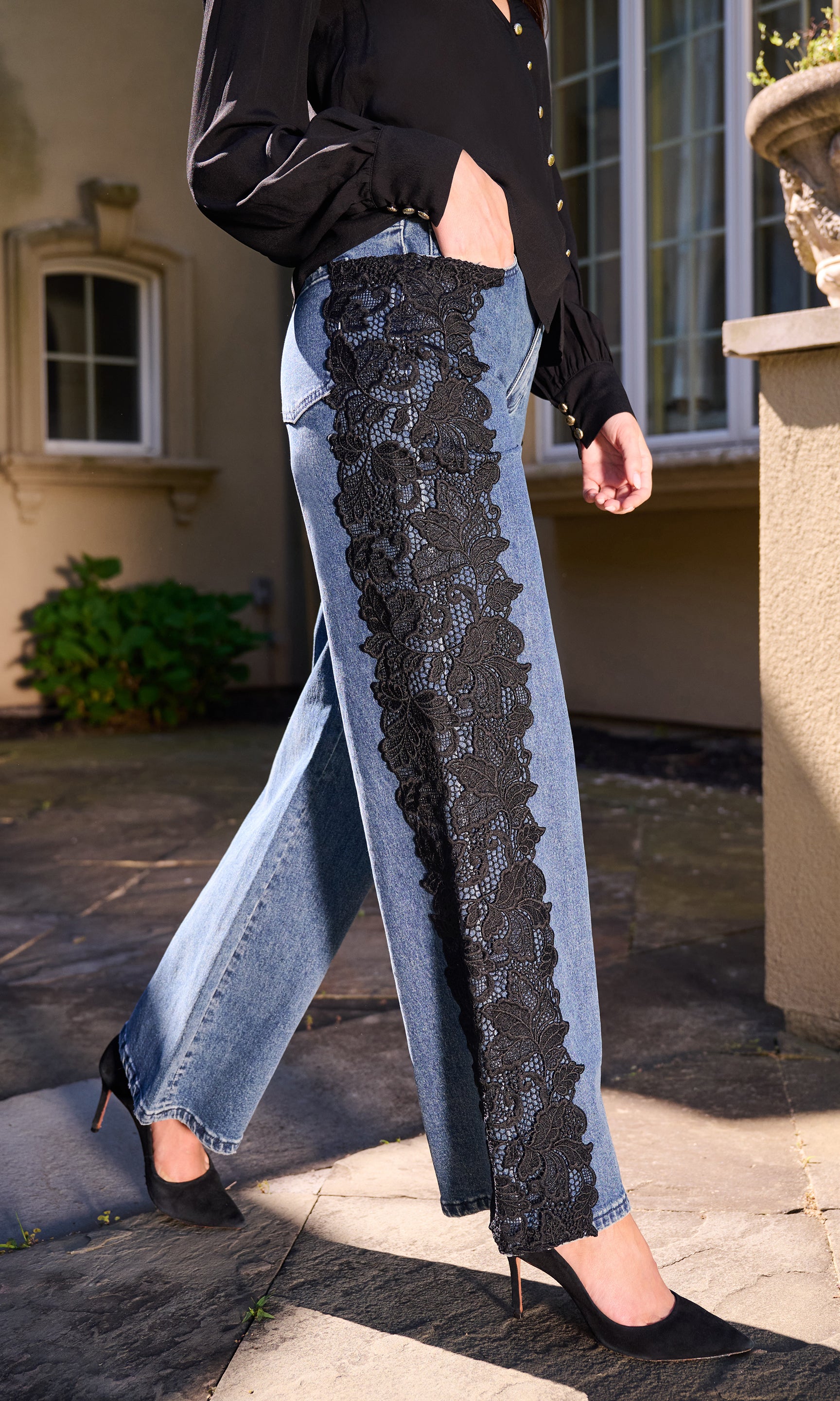 Model wearing light-wash wide-leg jeans with a prominent black lace panel down the front, styled with a black top, standing outdoors on a stone walkway.