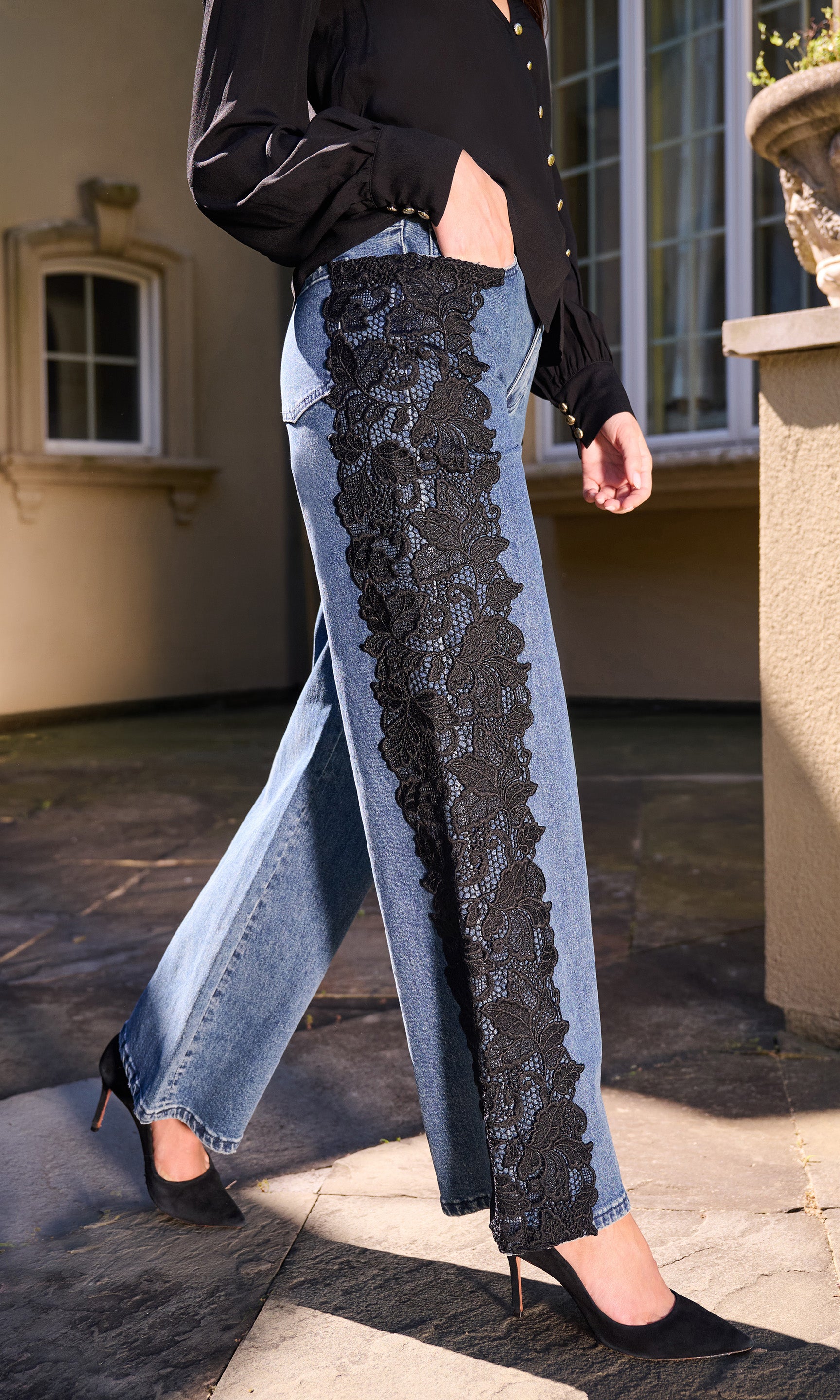 Model wearing light-wash wide-leg jeans with a prominent black lace panel down the front, styled with a black top, standing outdoors on a stone walkway.