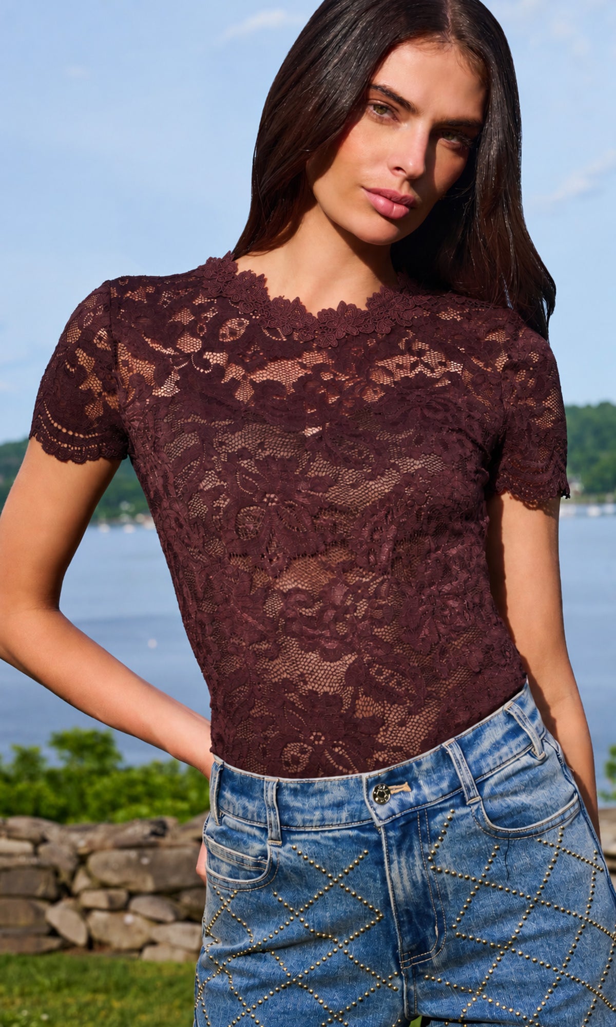 A woman wearing a short-sleeve burgundy lace top and blue jeans, standing outdoors with a lake and stone wall in the background.