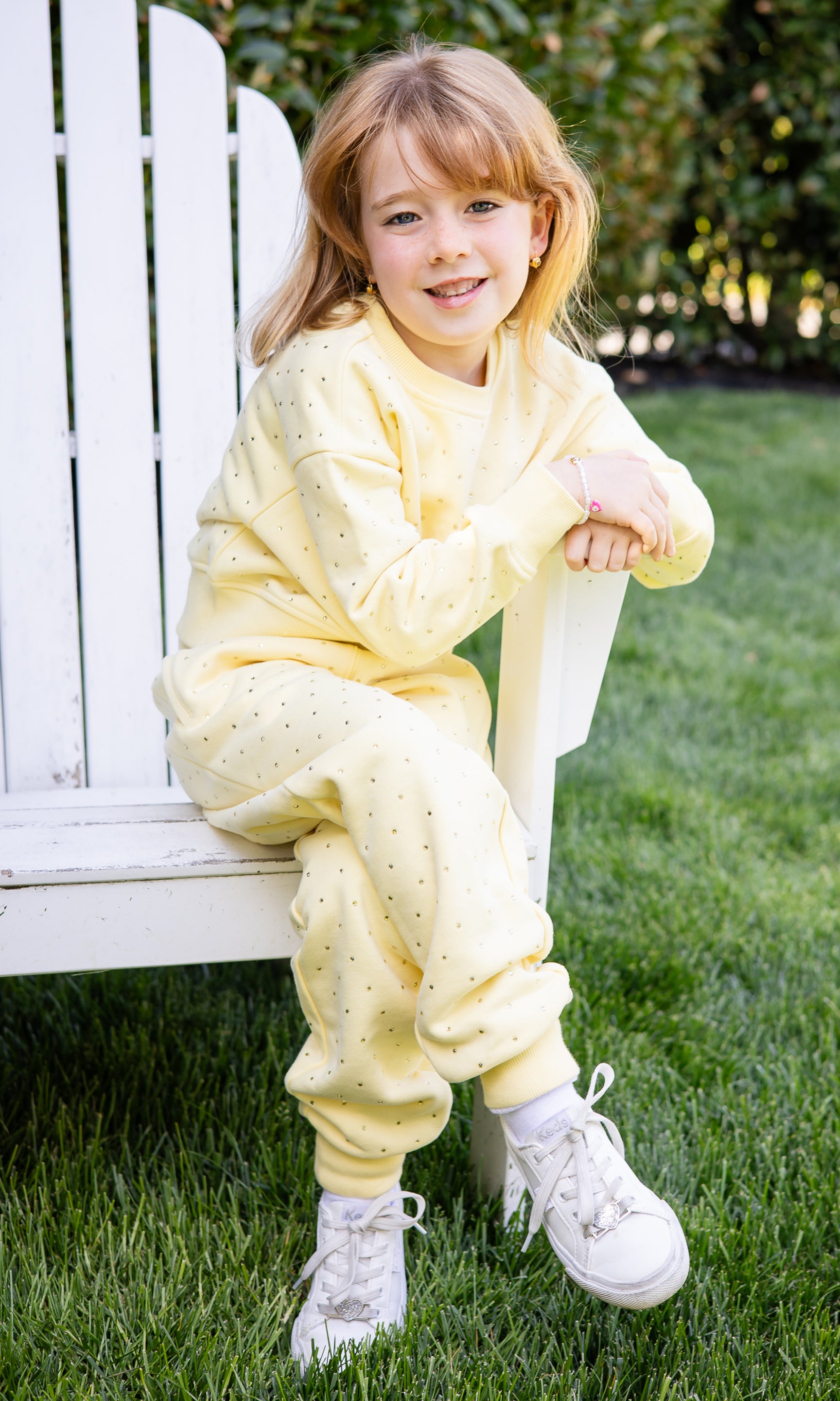 Child wearing a yellow sweatsuit sitting on a white chair outdoors.