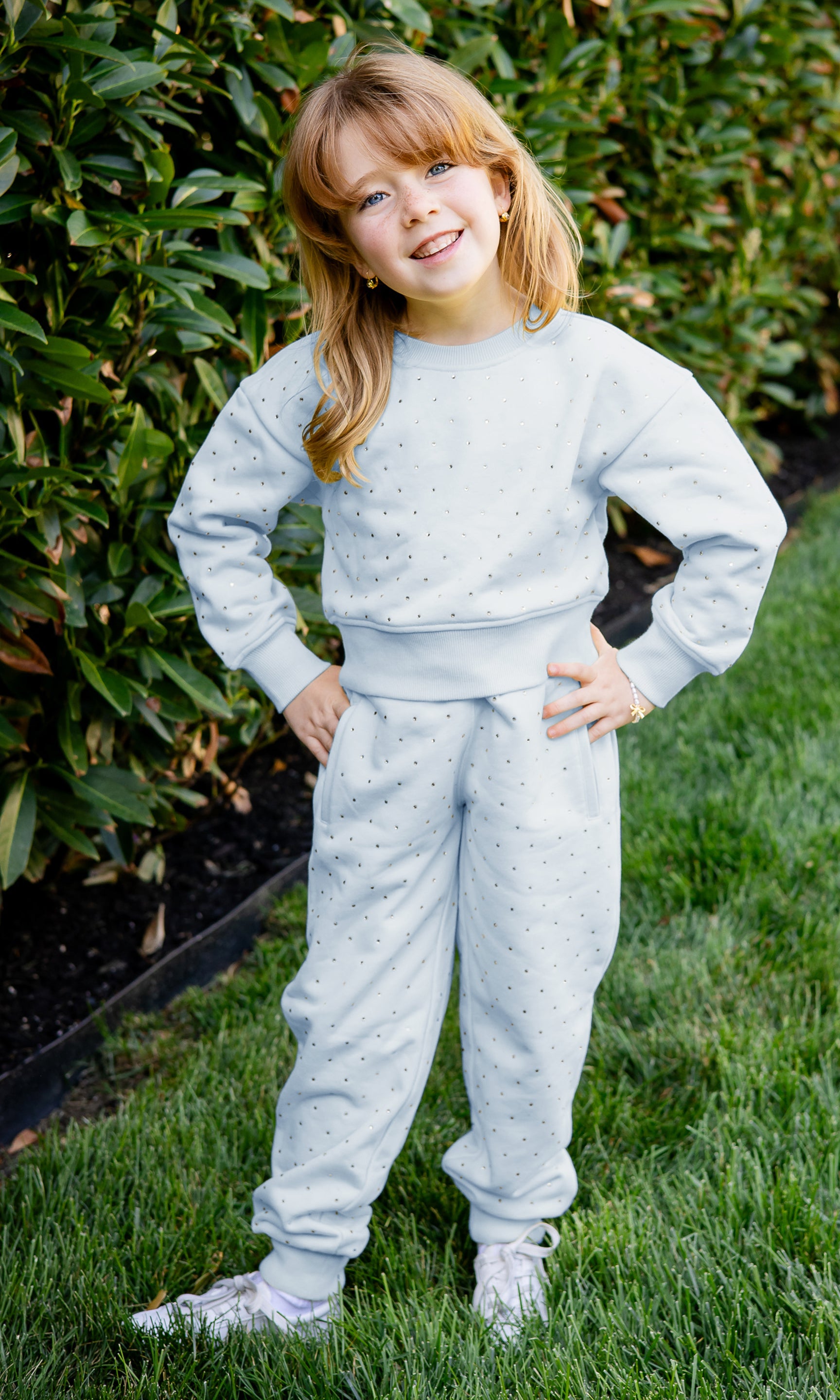 Child wearing a light blue outfit standing outdoors with greenery in the background
