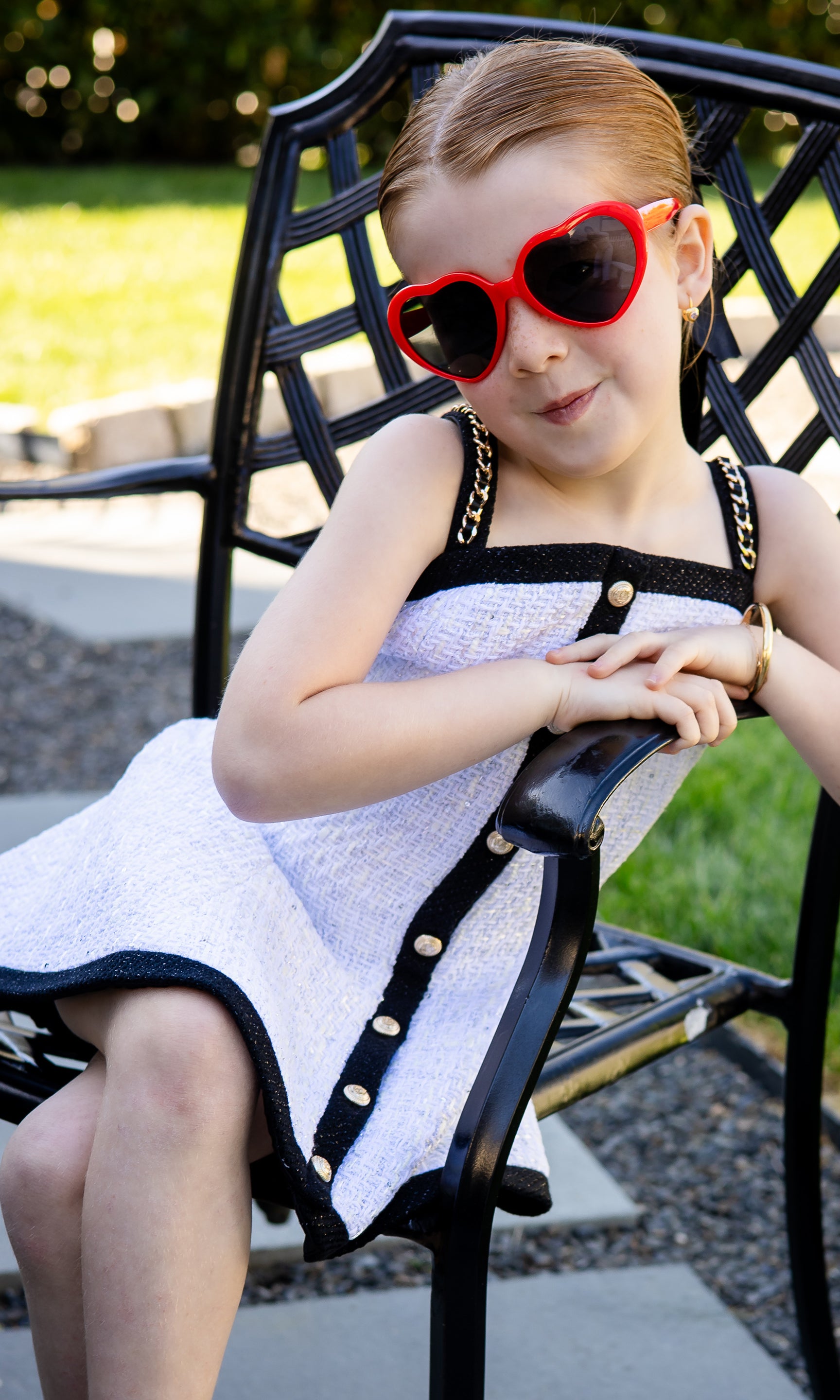 Child wearing a dress and heart-shaped sunglasses sitting on a chair outdoors.