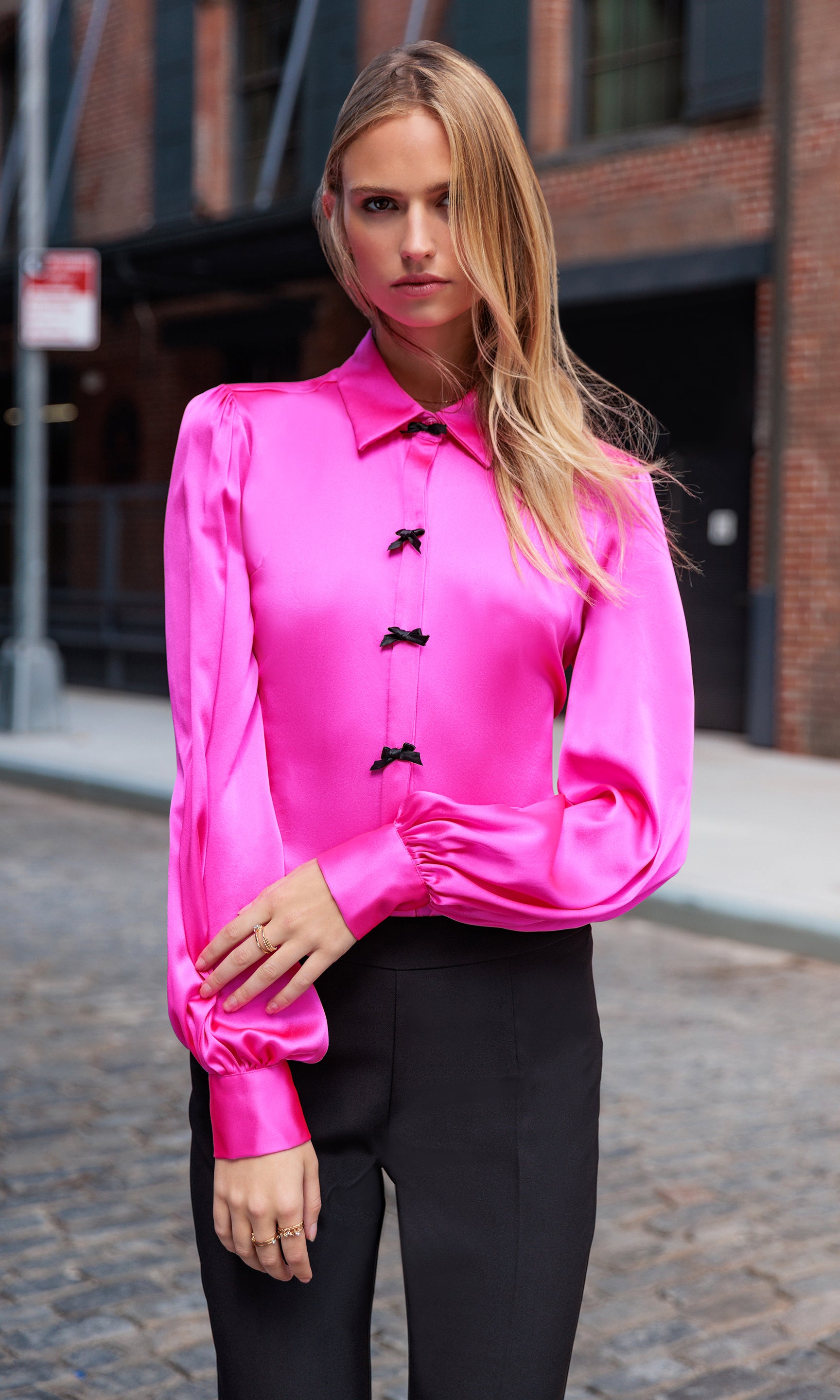 Woman wearing a bright pink blouse with black bow buttons on a city street.