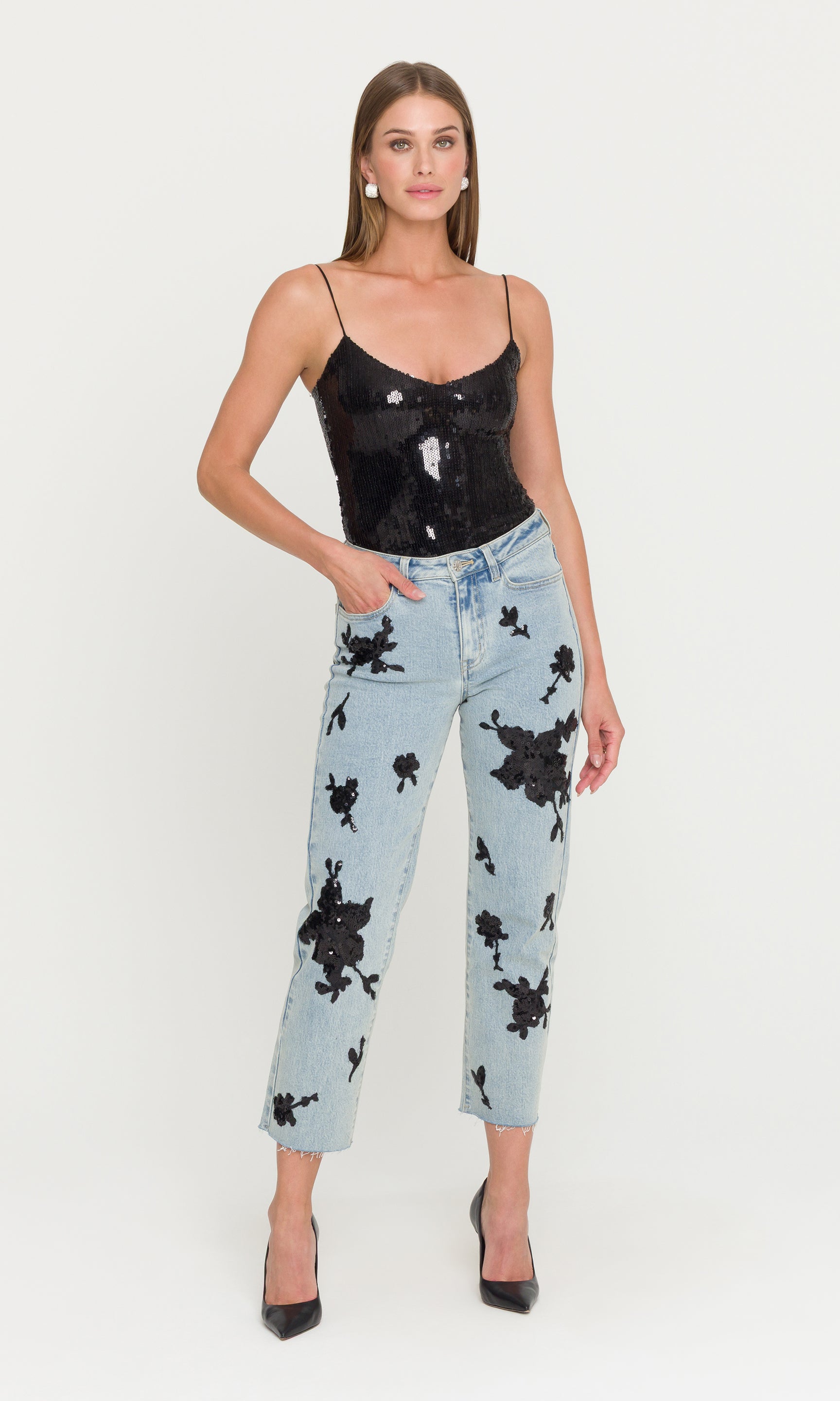 Full body image of model wearing a black sequin top and light blue jeans with black floral patterns on a white background