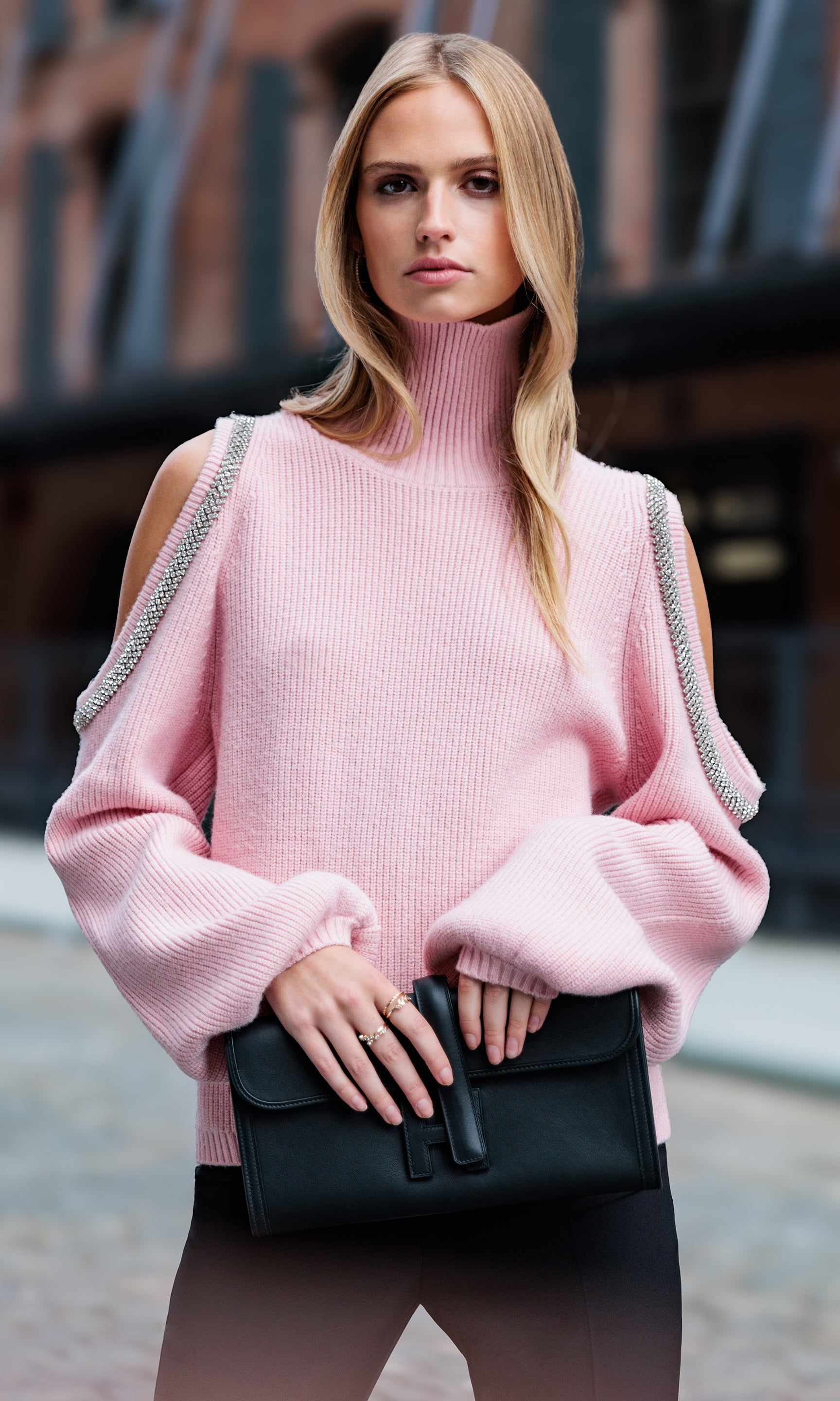 Woman wearing a pink sweater with cold-shoulder design, holding a black handbag.