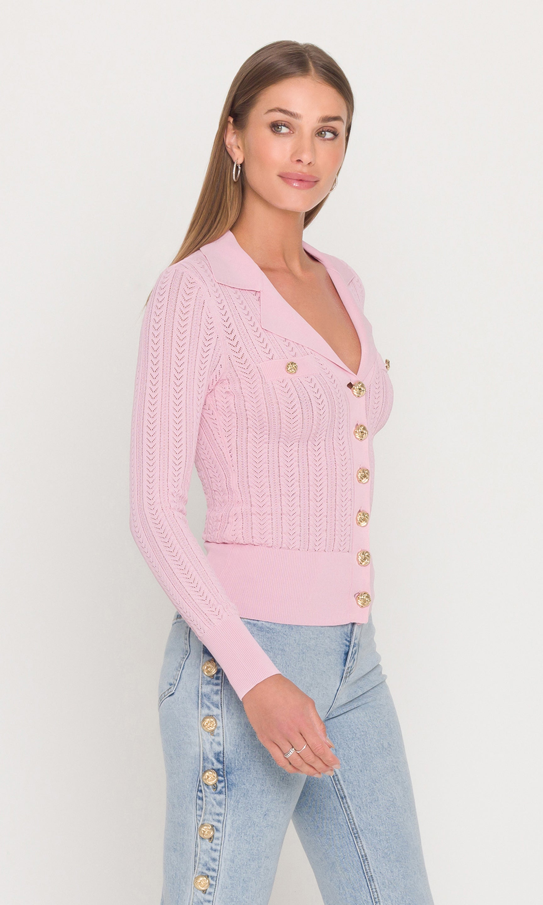 Side view of woman wearing a pink cardigan with gold buttons and light blue jeans on a white background