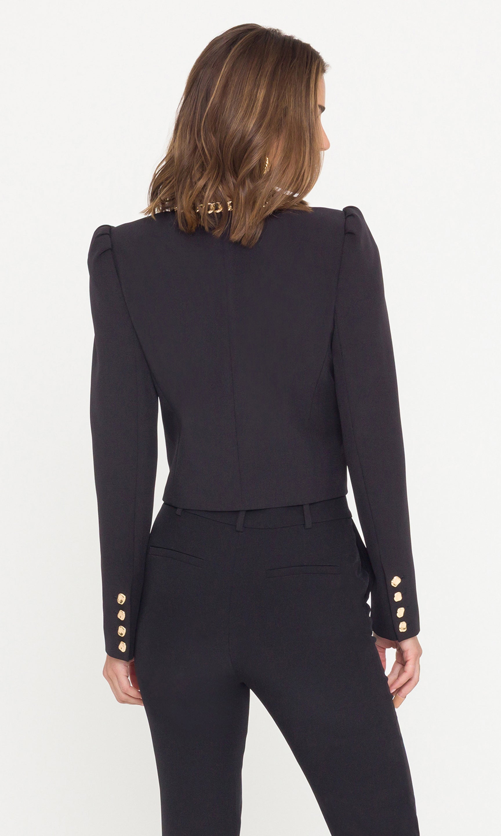 Back view of model wearing a black cropped jacket with a gold chain neckline and gold buttons on the sleeves