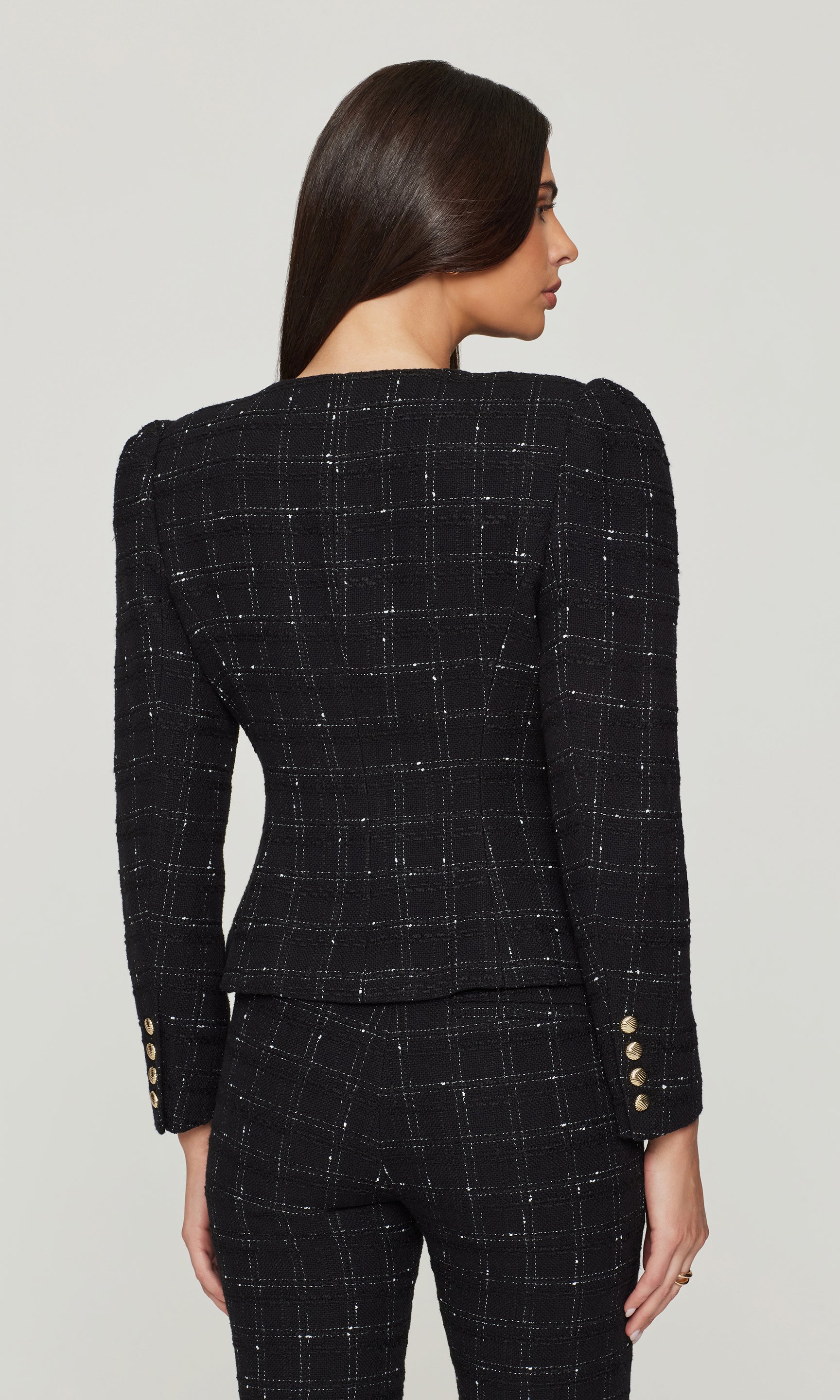 Back view of woman wearing a black checkered suit with gold buttons on a light gray background