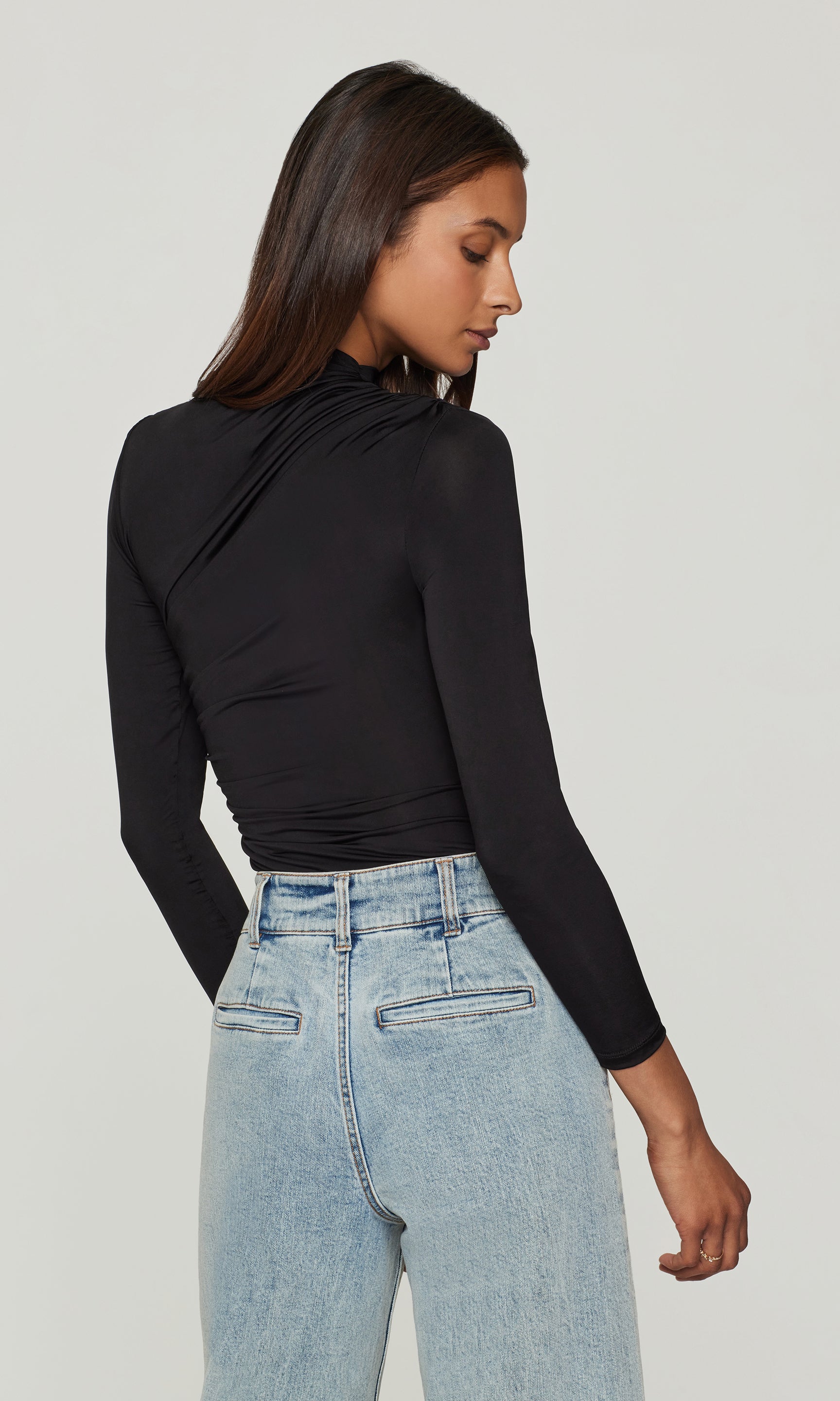 Back view of woman wearing a black long-sleeve top and light blue jeans on a plain background