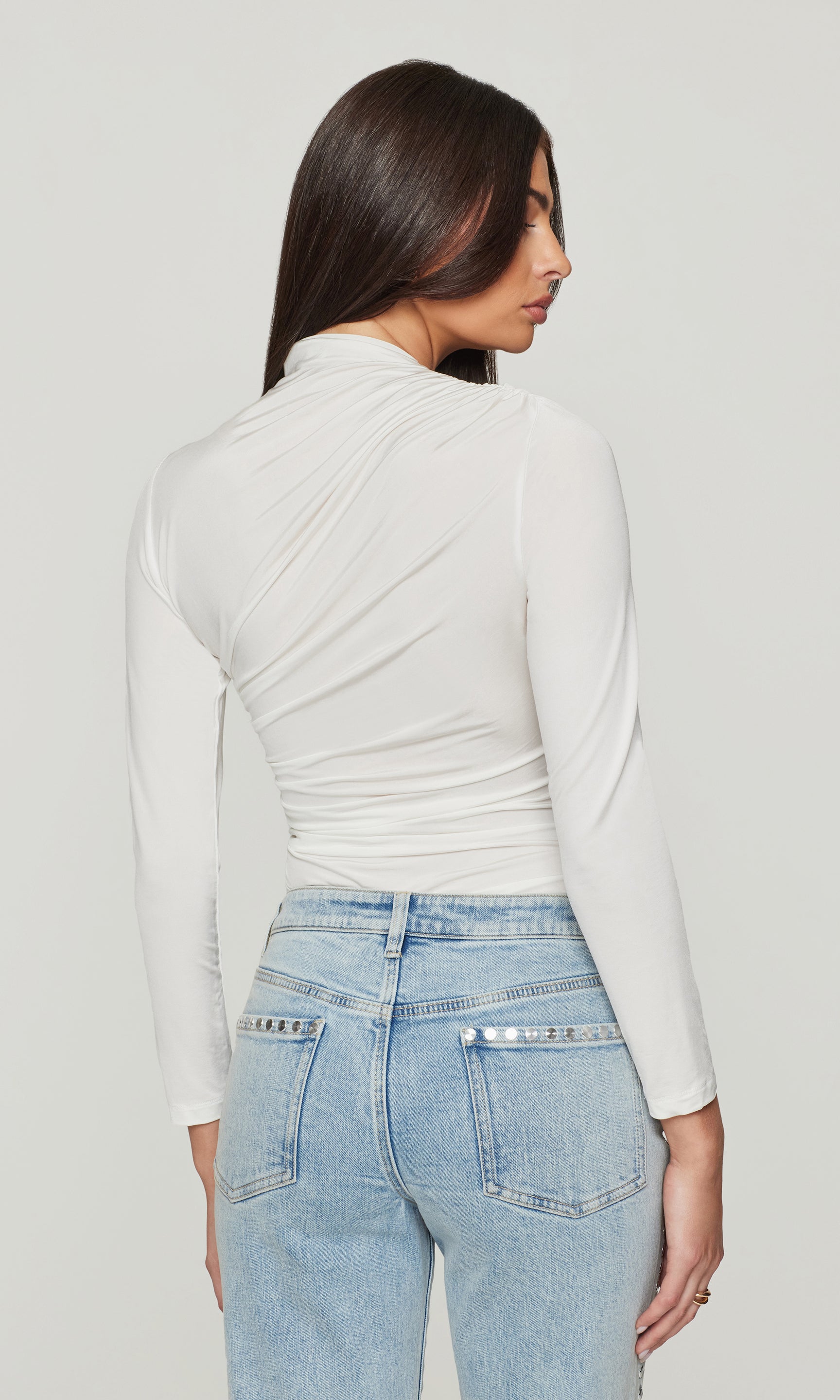 Back view of woman wearing a white long-sleeve top and light blue jeans on a plain background