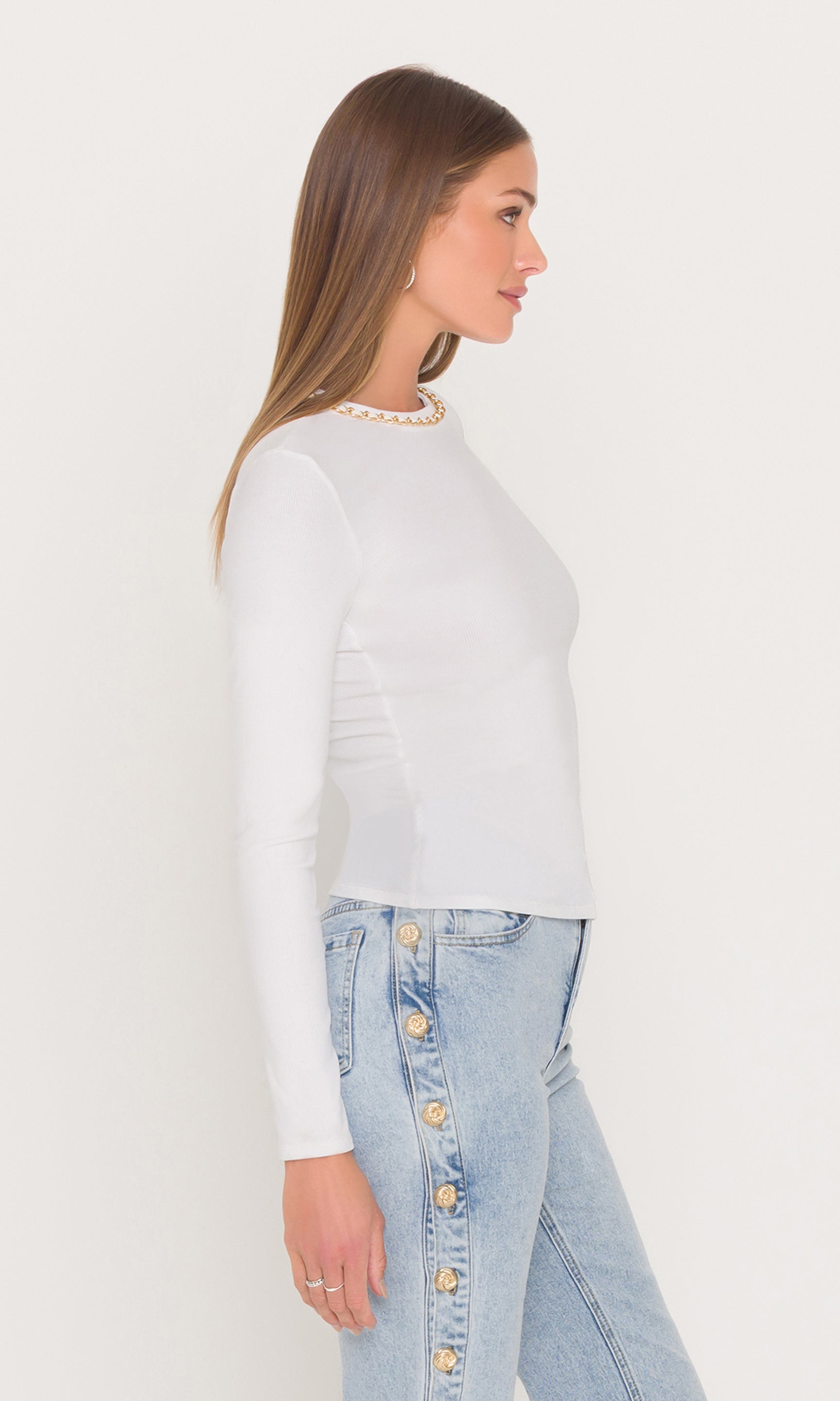 Side view of woman wearing a white long sleeve top with a gold chain neckline and blue jeans