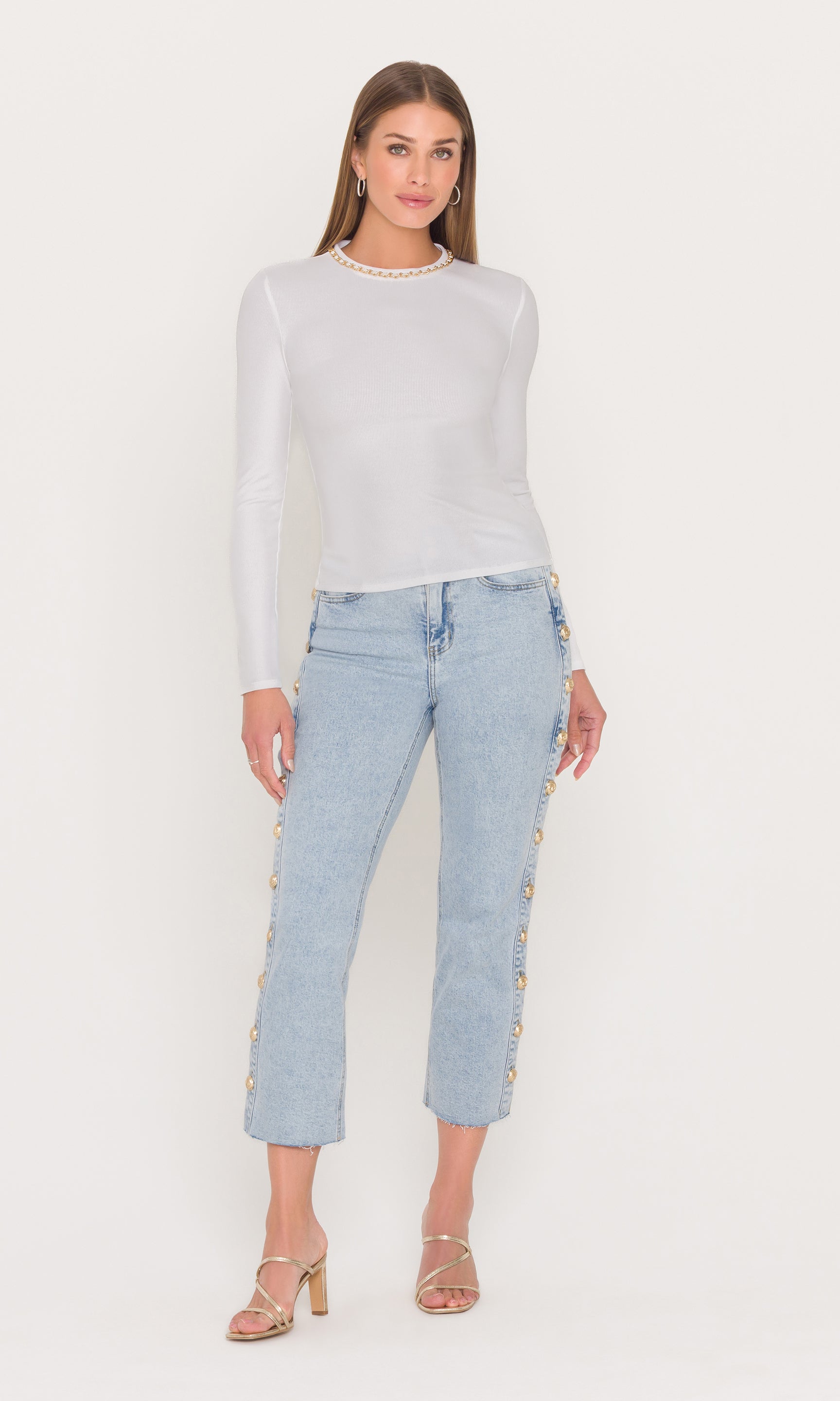 Full body image of model wearing a white long sleeve top and light blue jeans with decorative side leg buttons