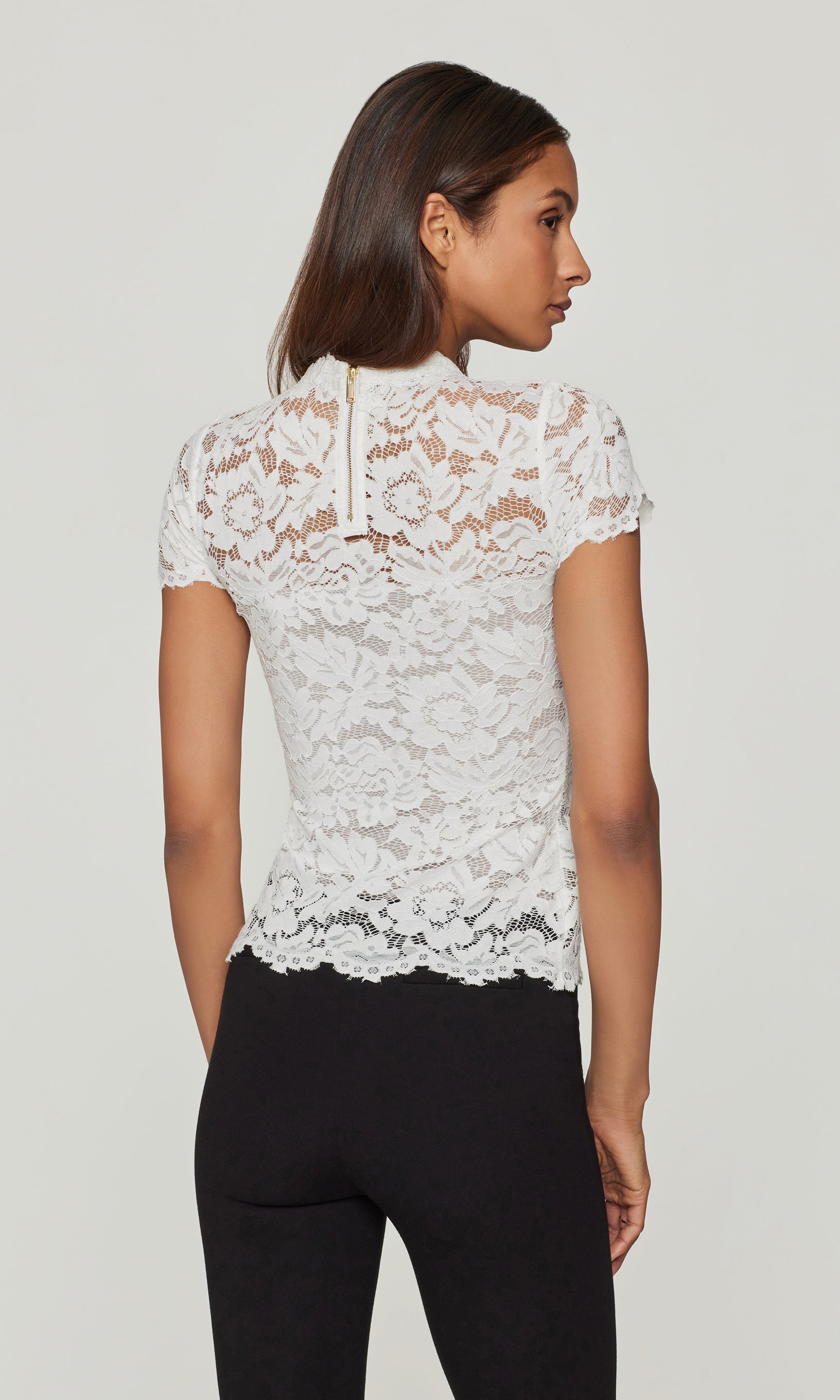 Back view of woman wearing a white lace top and black pants on a plain background