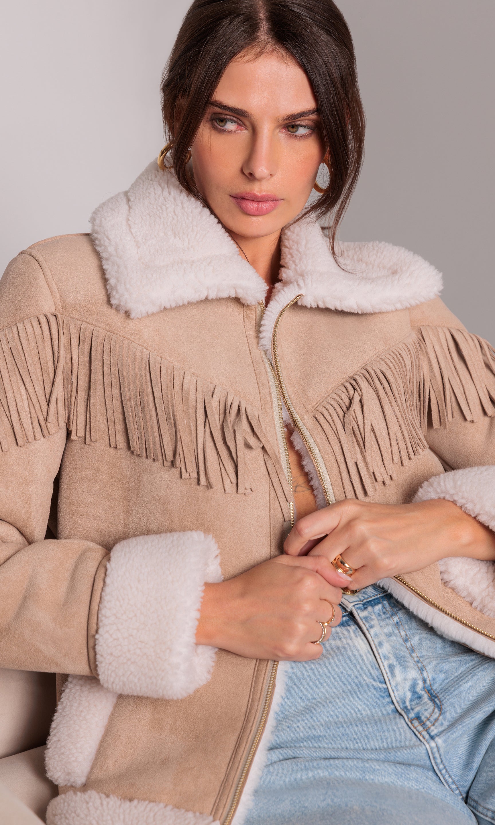 Woman wearing a beige shearling jacket with white fur trim and fringe details.