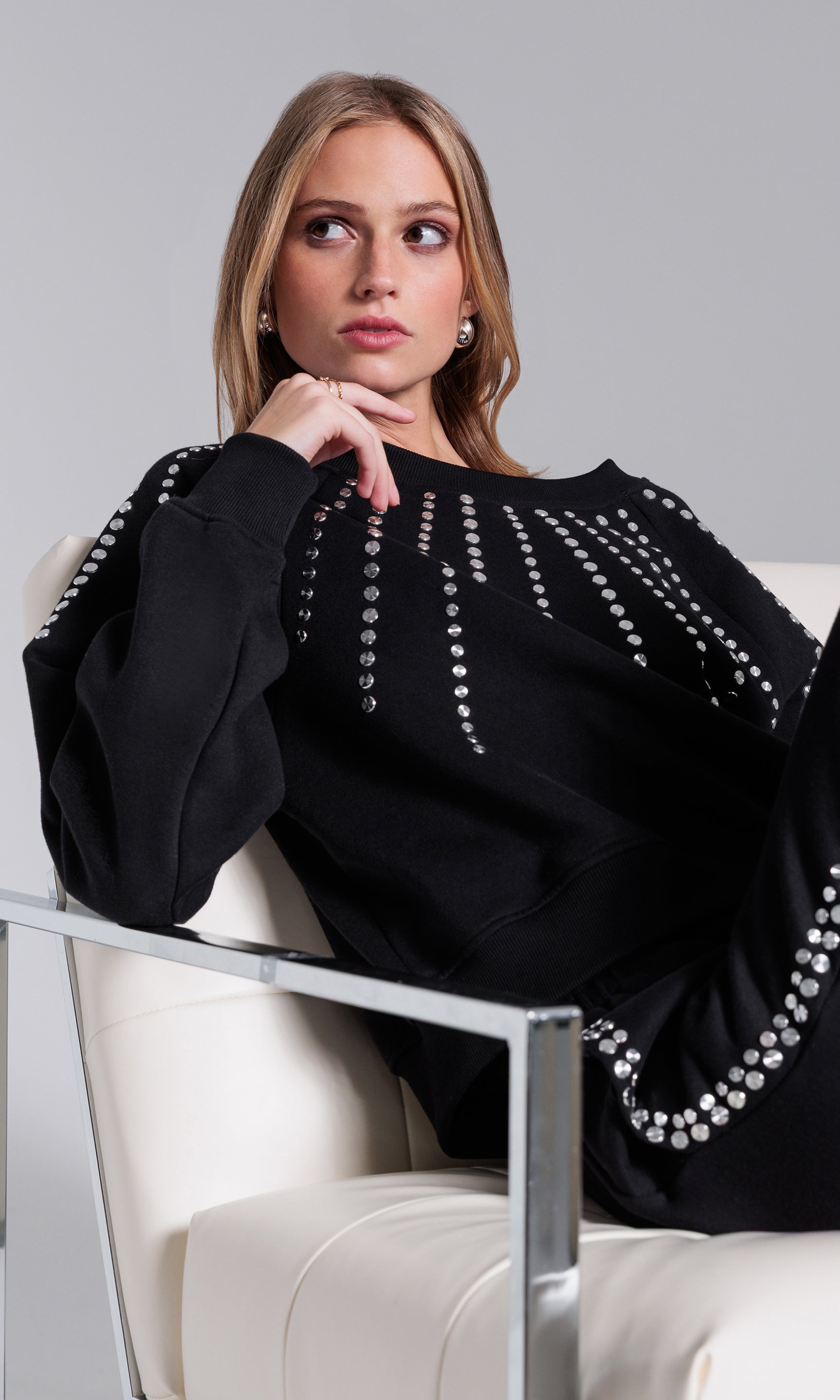 Woman wearing a black outfit with pearl embellishments sitting on a white chair against a gray background