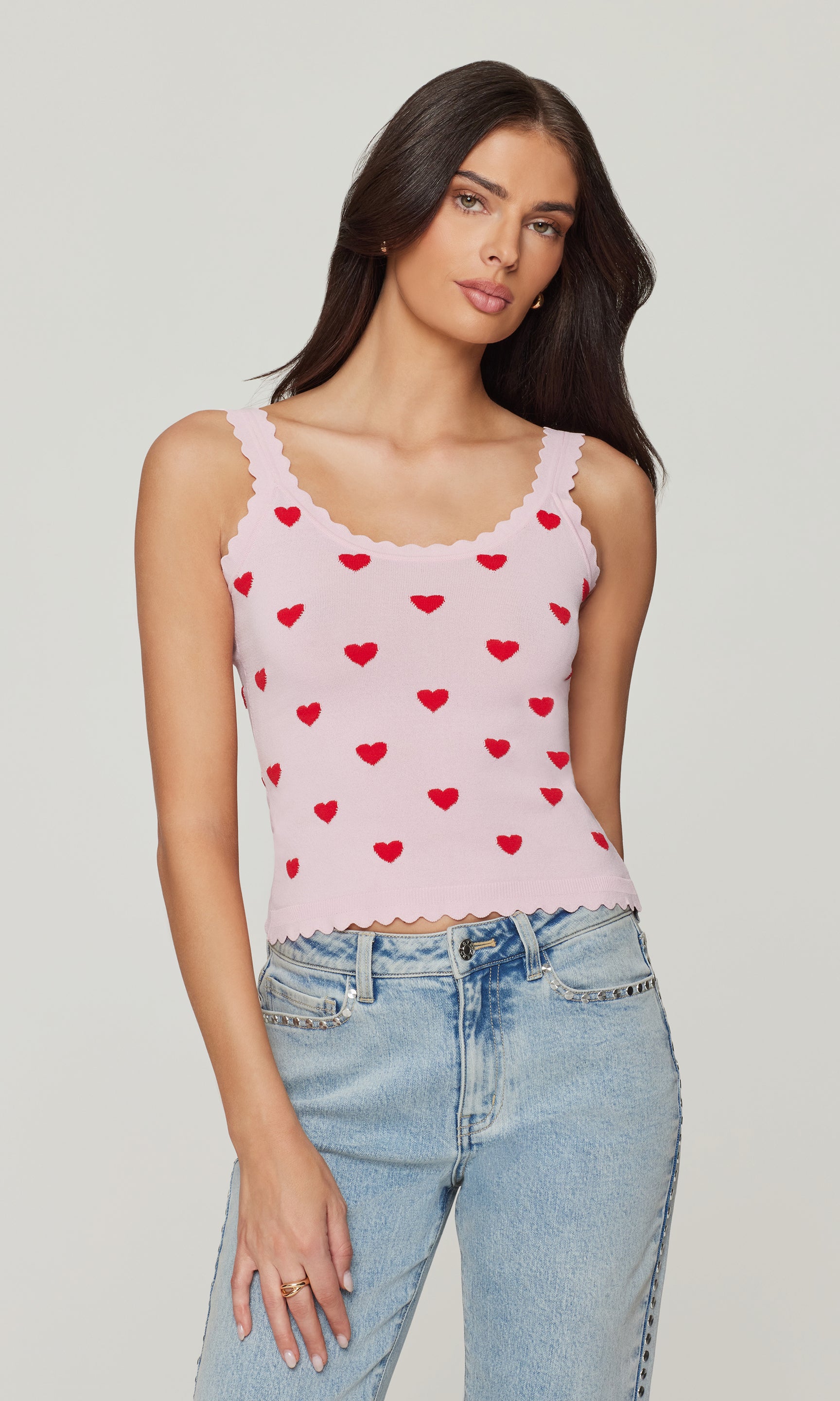 Woman wearing a pink tank top with red heart patterns and blue jeans on a plain background