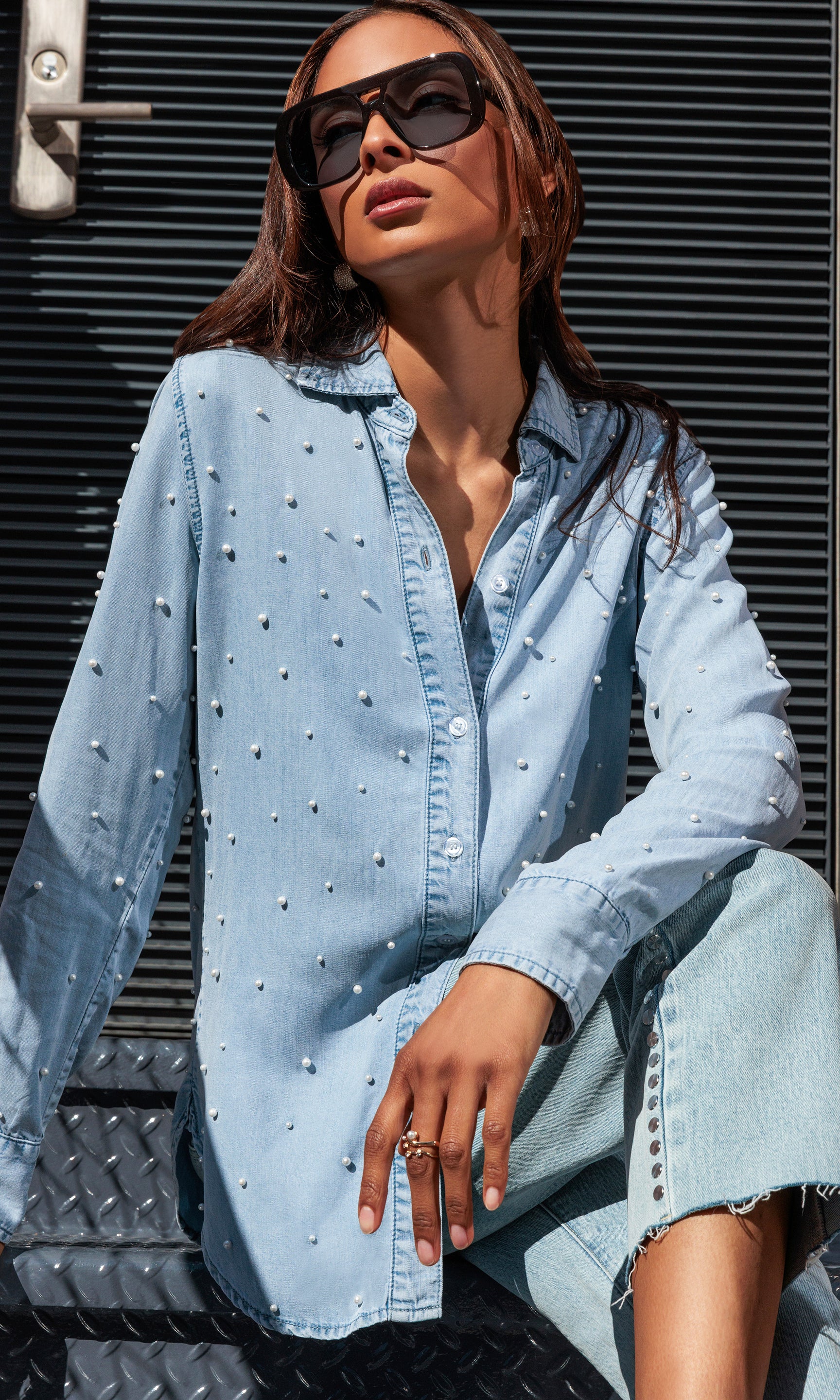 Model sitting on a door step wearing a light blue denim shirt with pearl embellishments and sunglasses