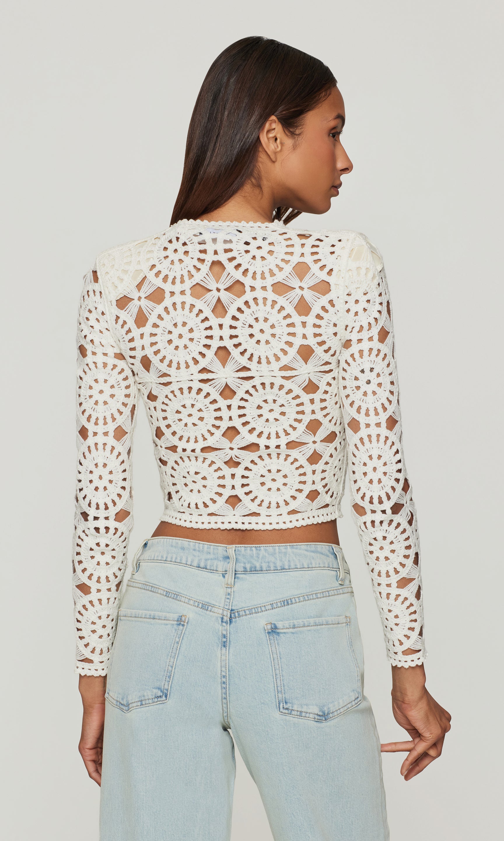 back view of model wearing a white crochet long-sleeve top and light blue jeans on a plain background
