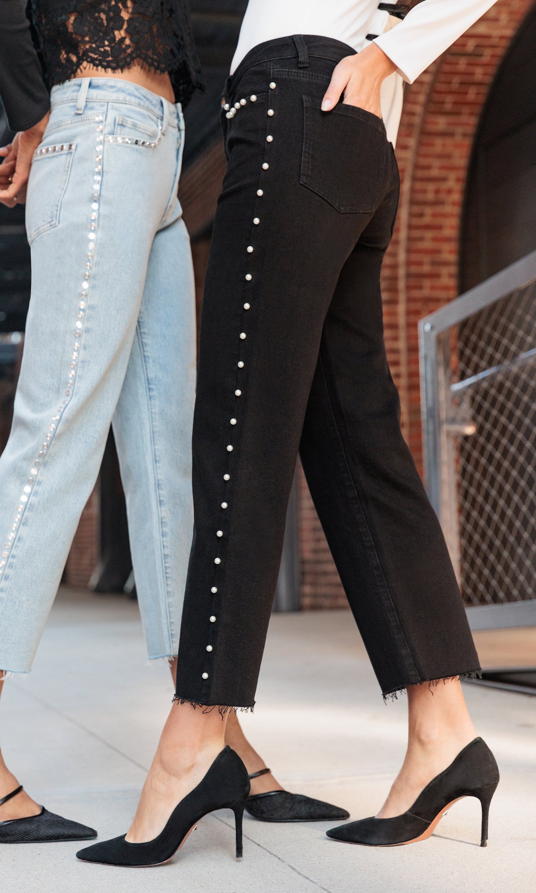 Person wearing black pants with white pearls and light blue jeans, standing on a sidewalk.
