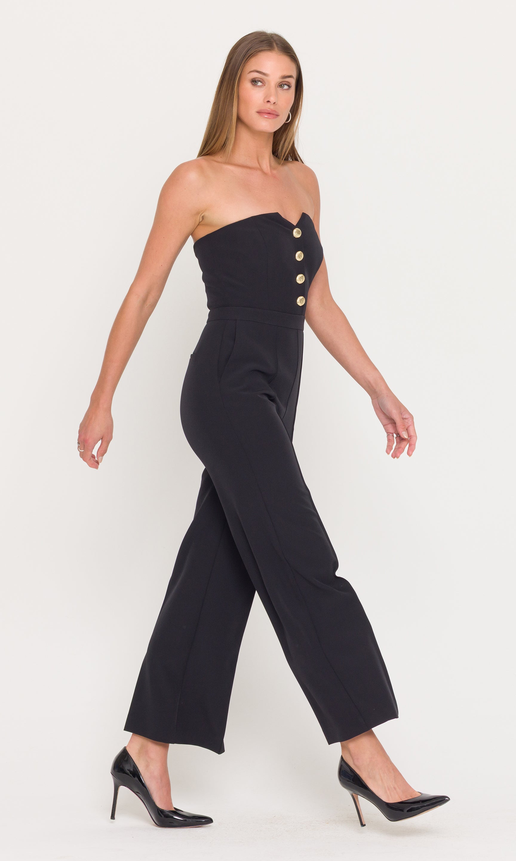 Side view of woman wearing a black strapless jumpsuit with gold buttons on a white background