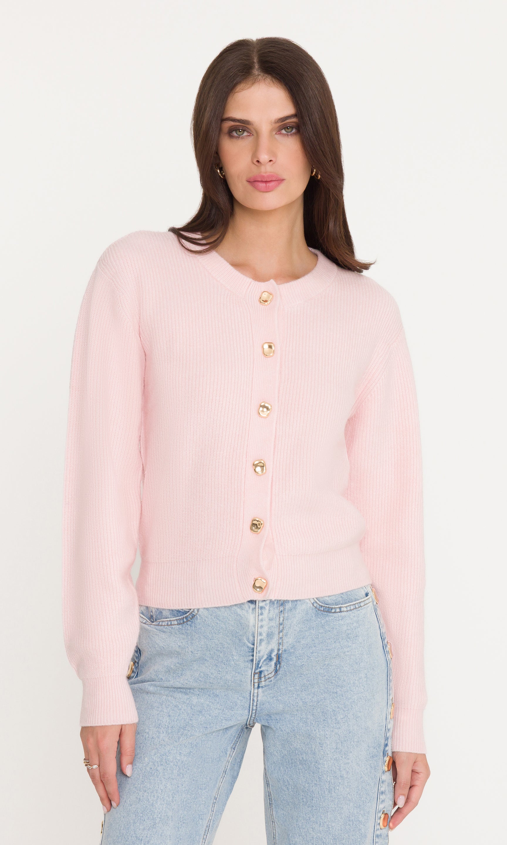 Woman wearing a light pink cardigan with gold buttons and light blue jeans on a white background