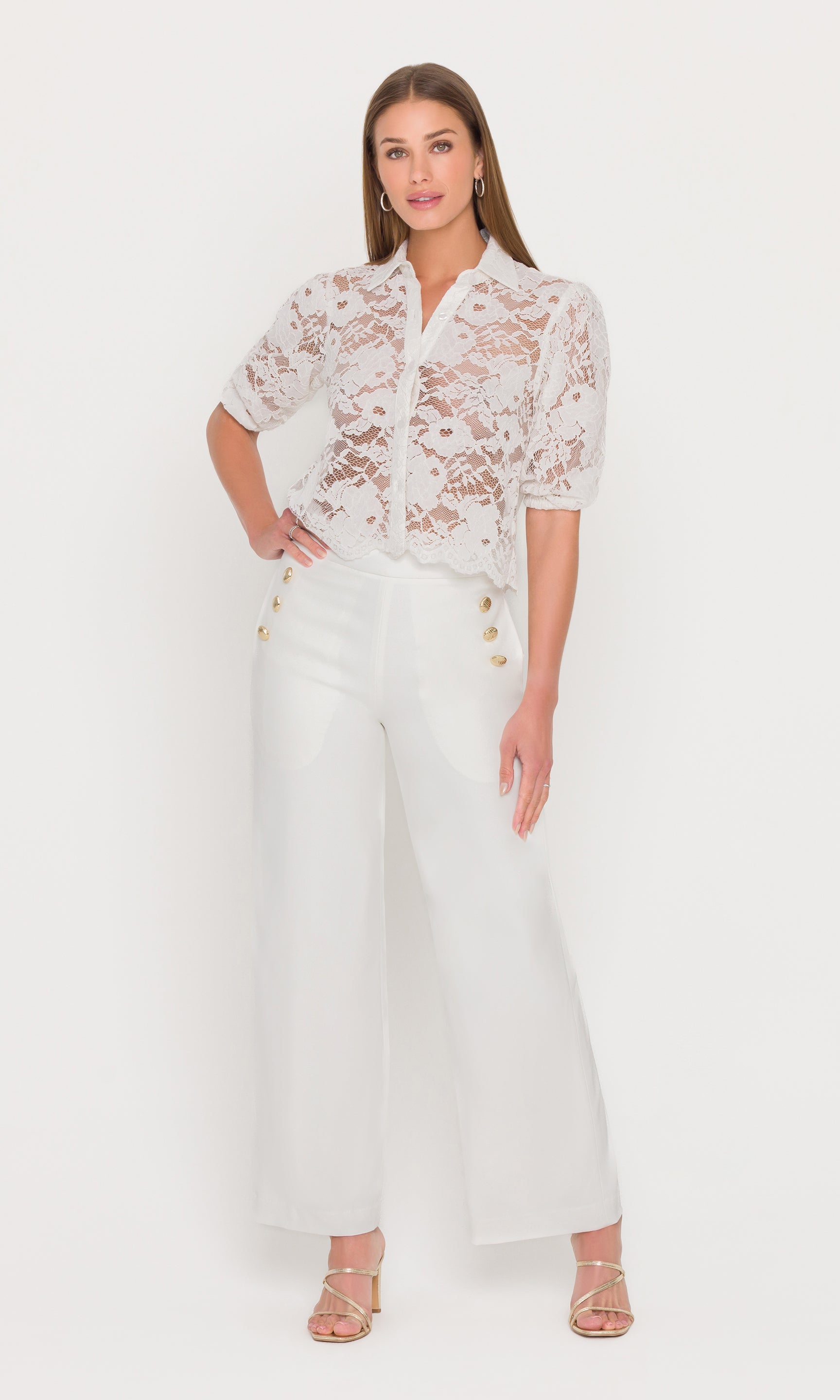 Full body image of model wearing a white lace blouse and white pants with gold button details on a plain background