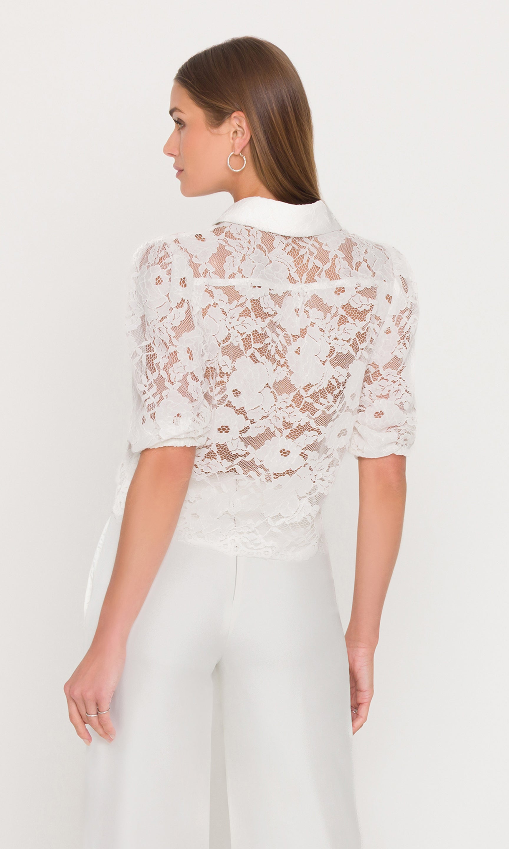 Back view of model wearing a white lace blouse and white pants with gold button details on a plain background