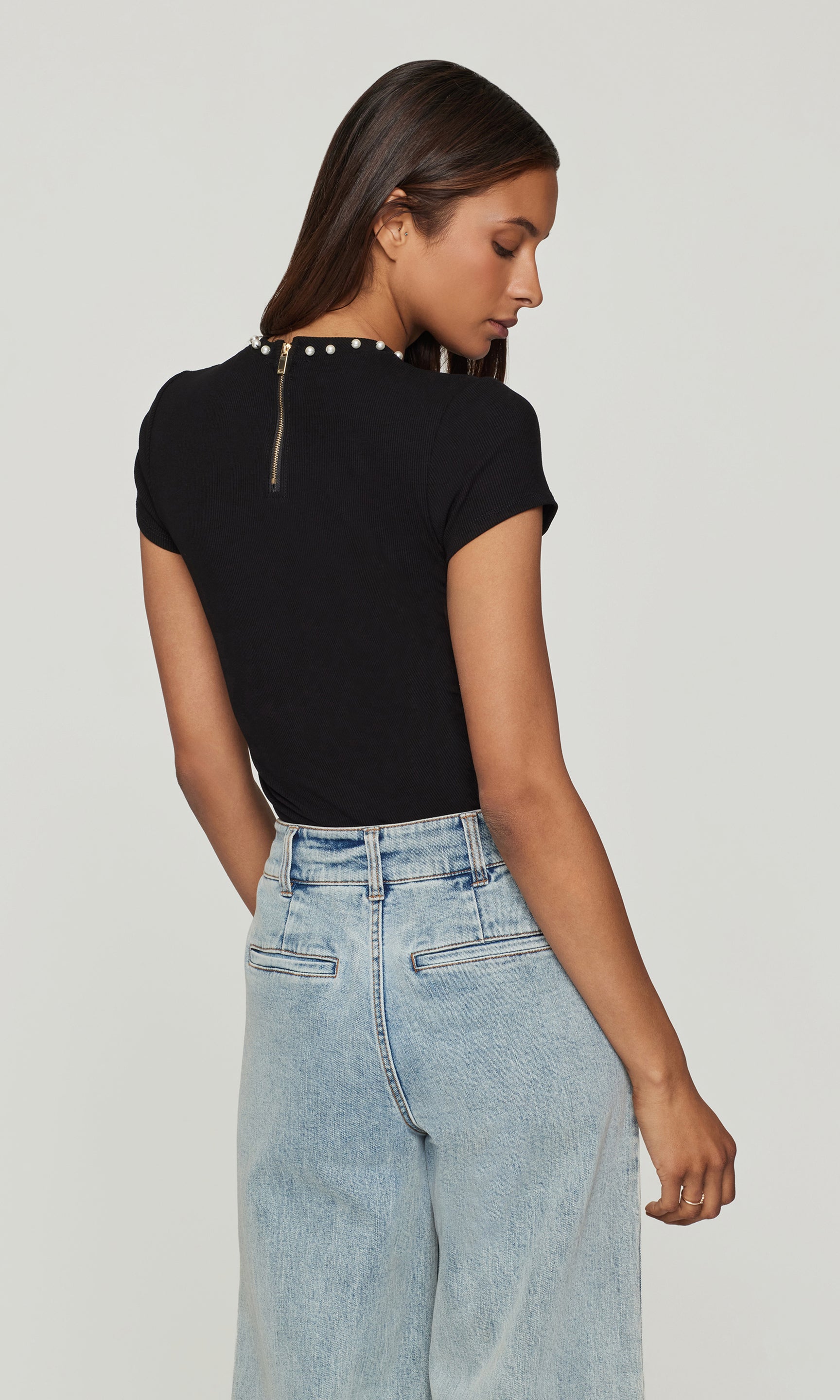 Back view of woman wearing a black top with studded details and light blue jeans on a plain background