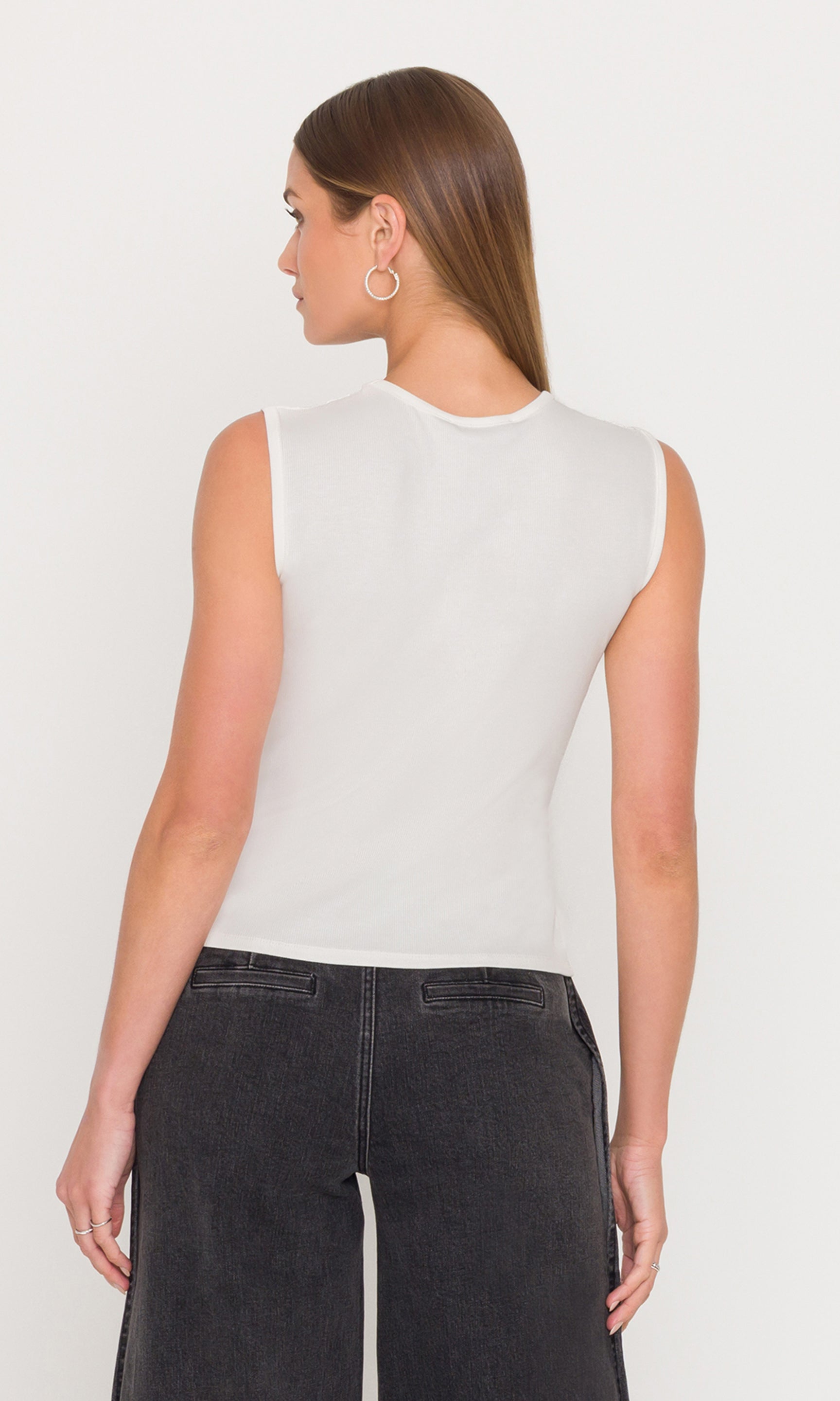 Back view of woman wearing a white sleeveless top and black pants on a plain background