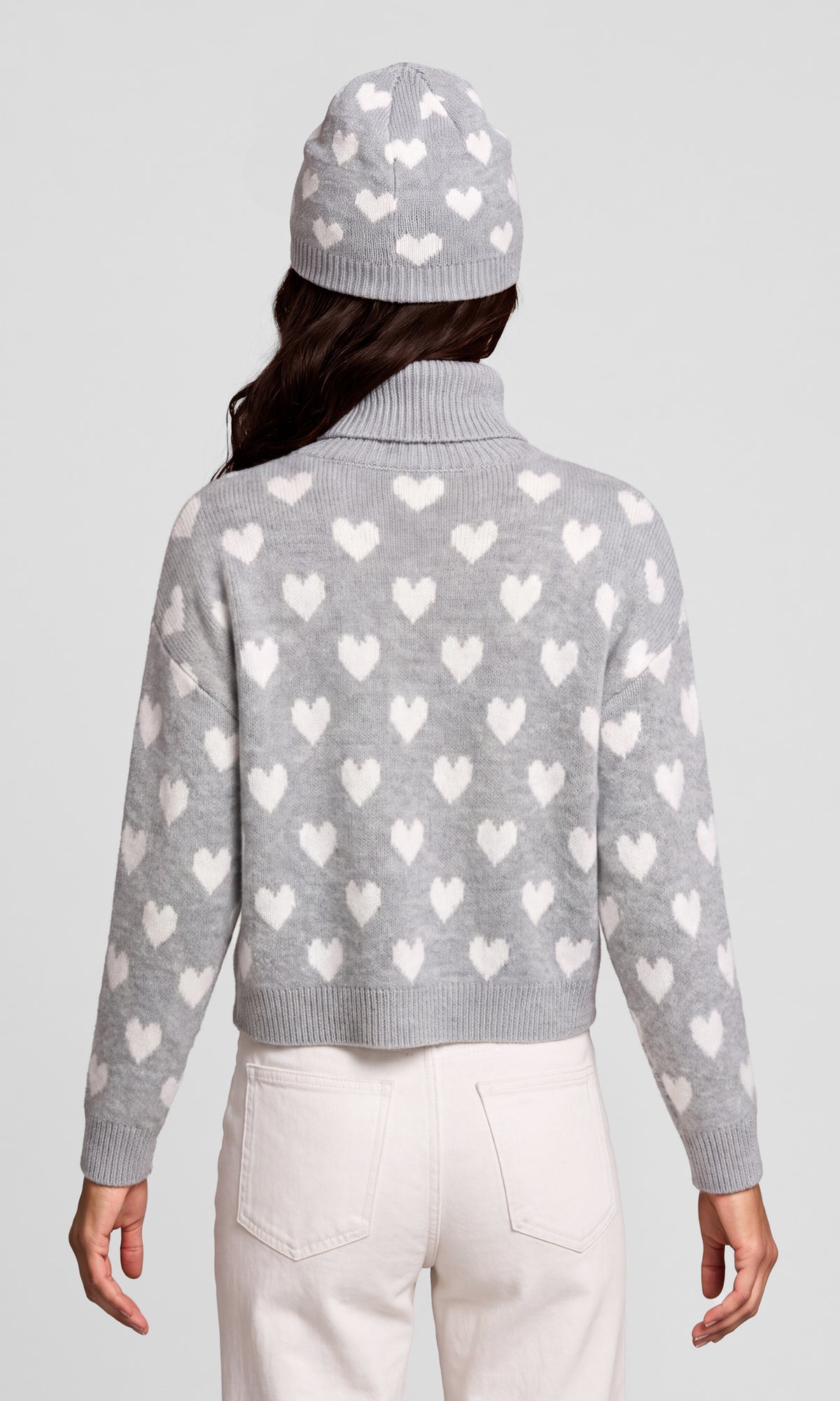 Back view of model wearing a light gray knit sweater with a repeating white heart pattern, a matching gray knit beanie, and off-white pants.