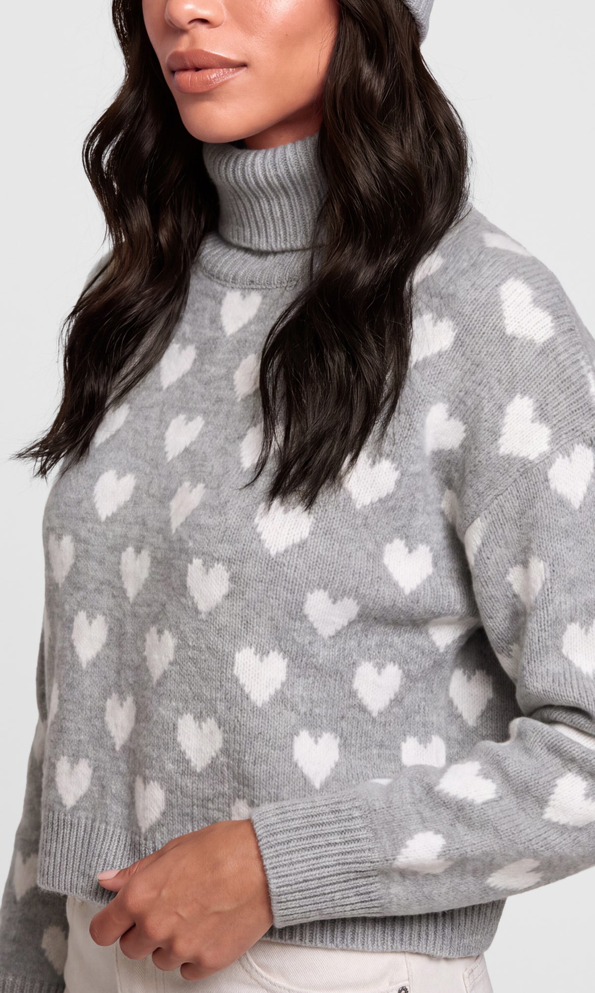 A close-up of a woman wearing a gray knit turtleneck sweater with a white heart pattern, paired with white pants.