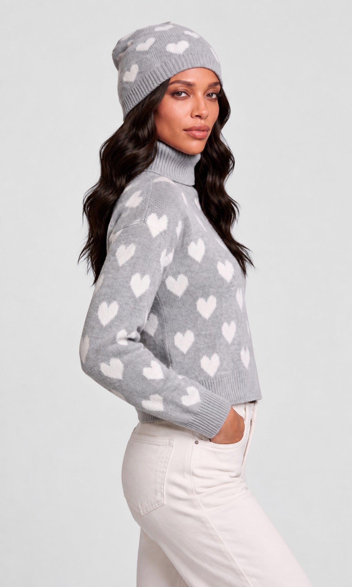 Side view of model wearing a light gray knit turtleneck sweater with a white heart pattern, a matching gray beanie, and off-white pants