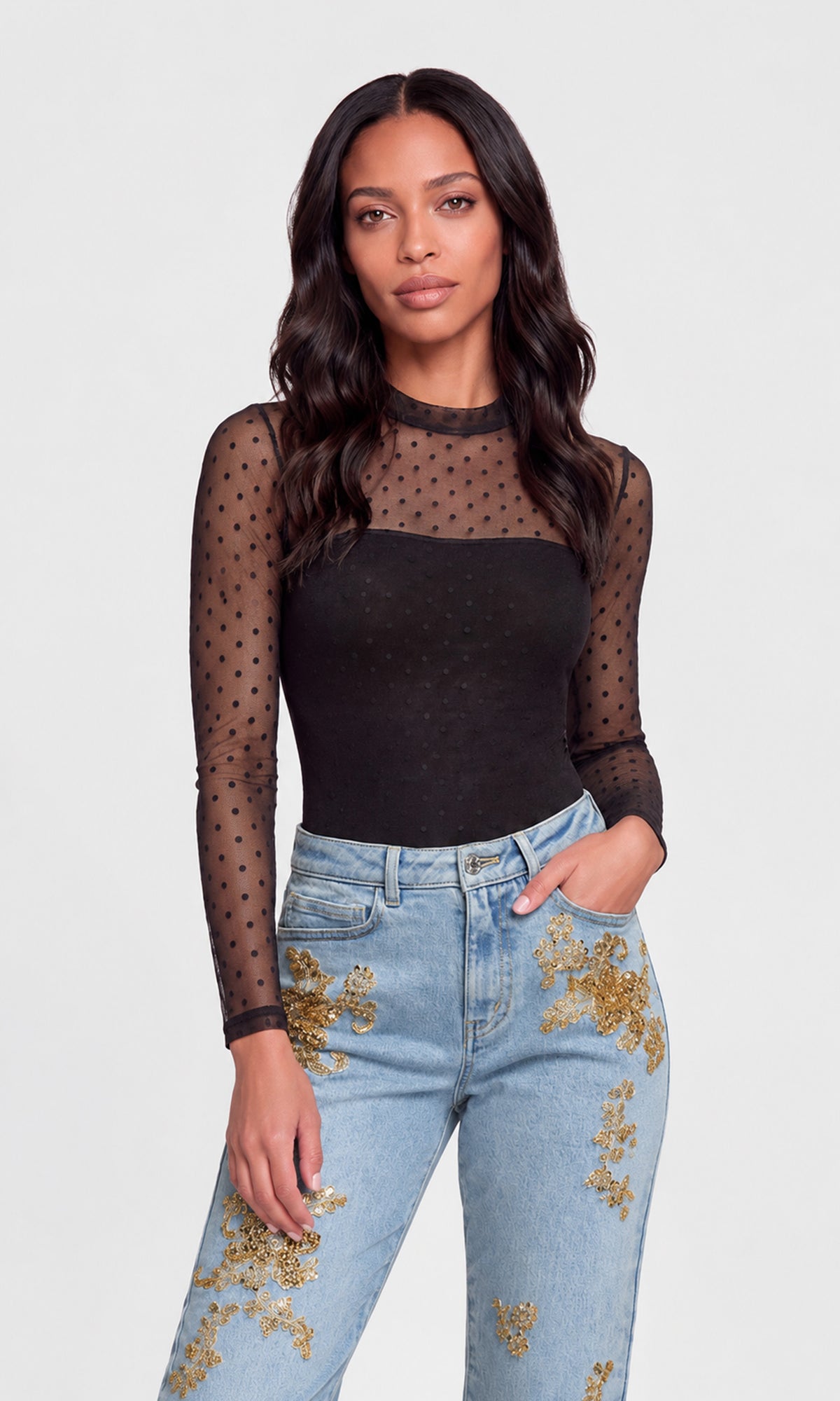 A woman modeling a black long-sleeve top with sheer black polka-dot mesh sleeves and a solid black square neckline, paired with light blue jeans.