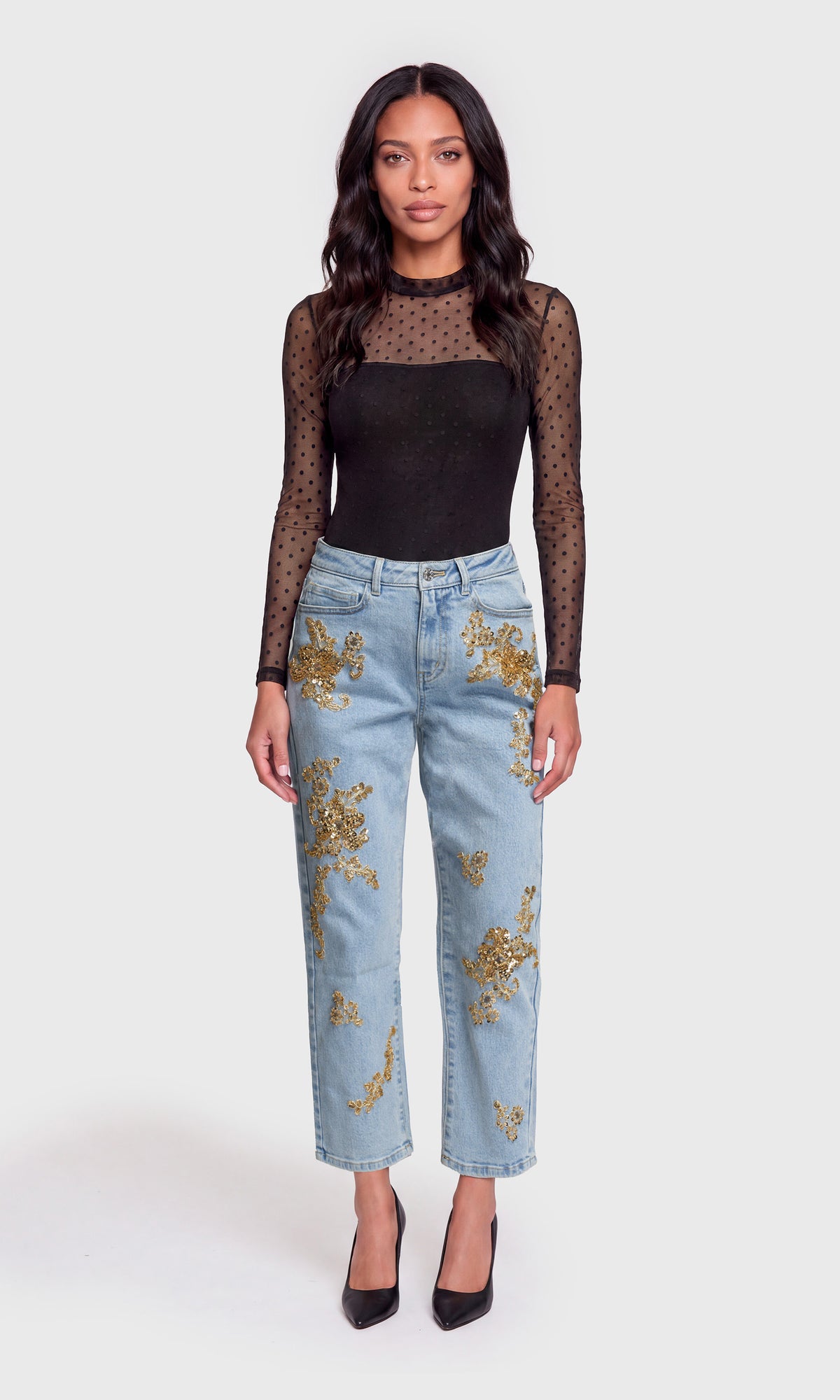 Model wearing a black sheer long-sleeve top, light-wash blue jeans with gold floral embroidery, and black pointed heels.