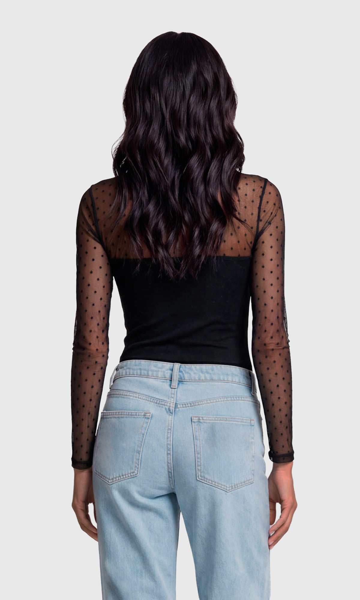 Back view of model wearing a black long-sleeve top with sheer black polka-dot mesh sleeves and a solid black square neckline, paired with light blue jeans.