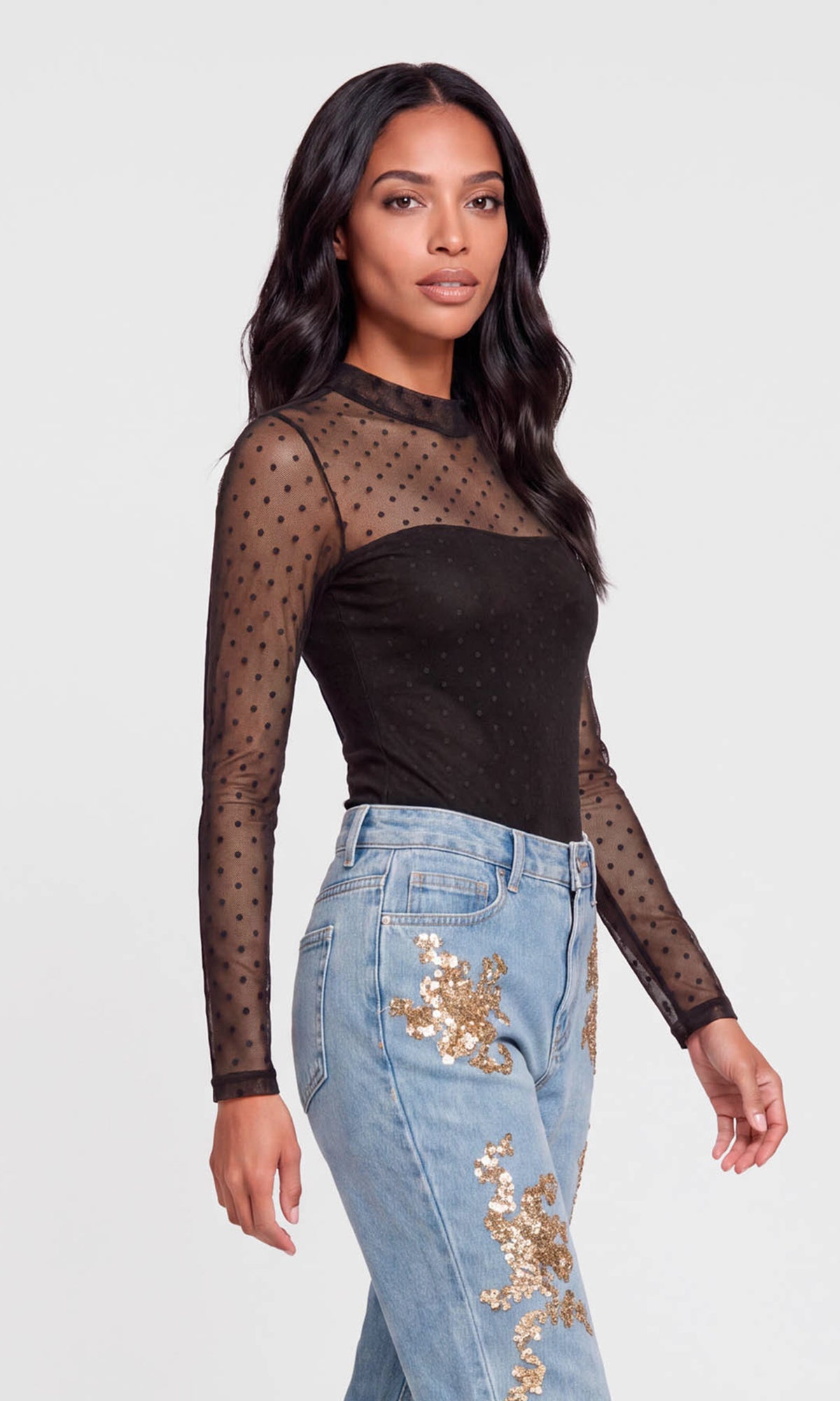 Side view of model wearing a black long-sleeve top with sheer black polka-dot mesh sleeves and a solid black square neckline, paired with light blue jeans.