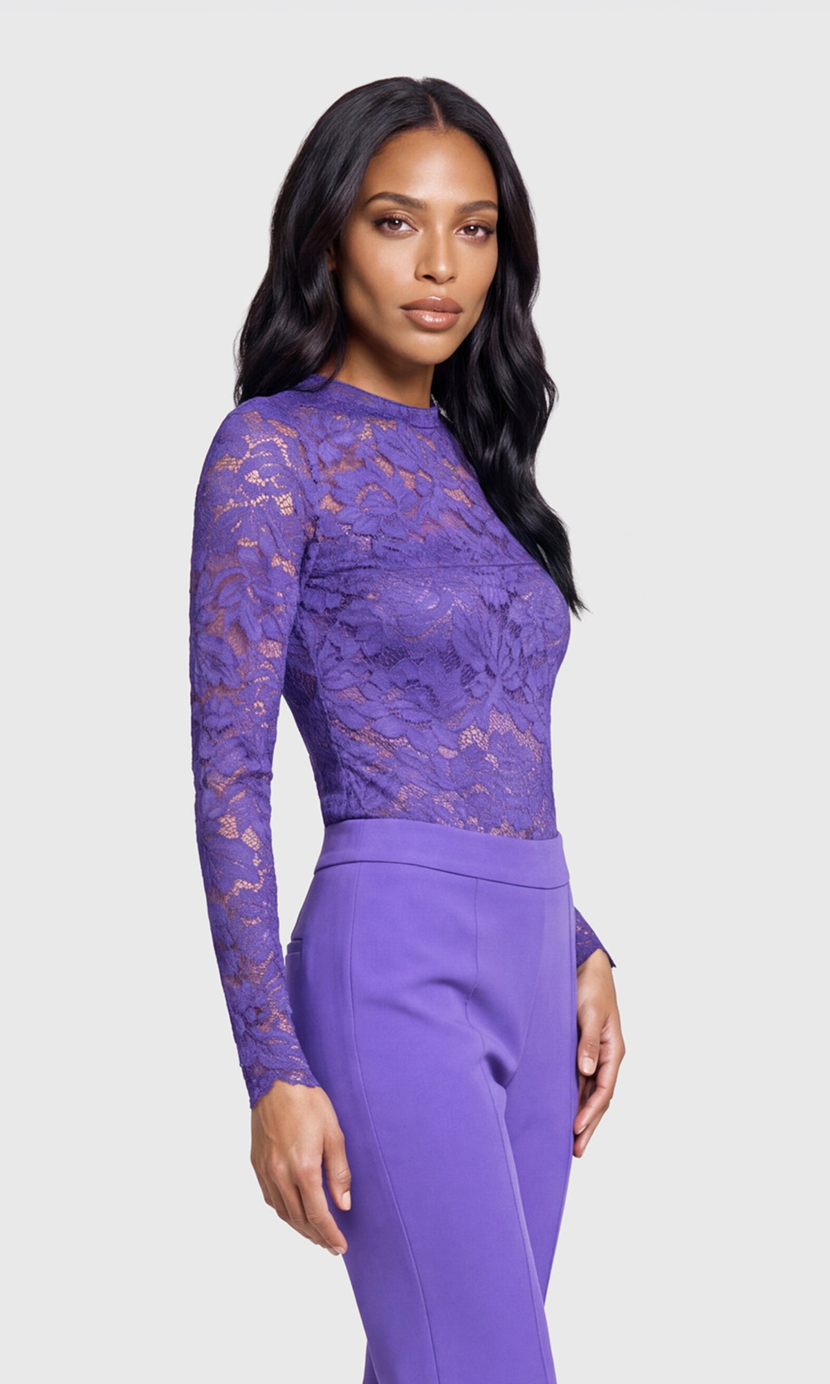 A woman modeling a purple long-sleeve lace top paired with matching purple trousers, photographed against a light gray studio background.