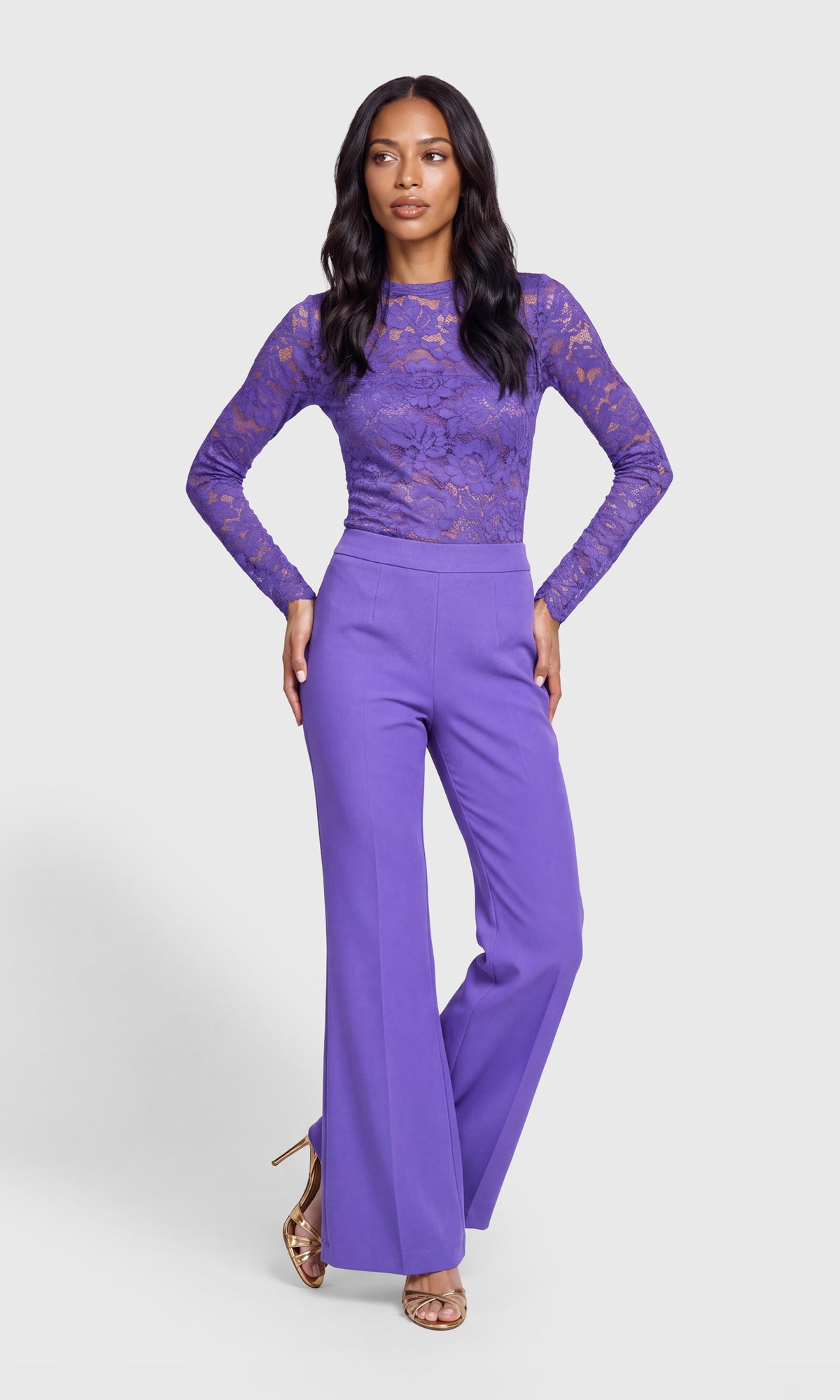 A woman modeling a purple long-sleeve jumpsuit with a lace bodice and wide-leg pants, standing against a light gray studio background.