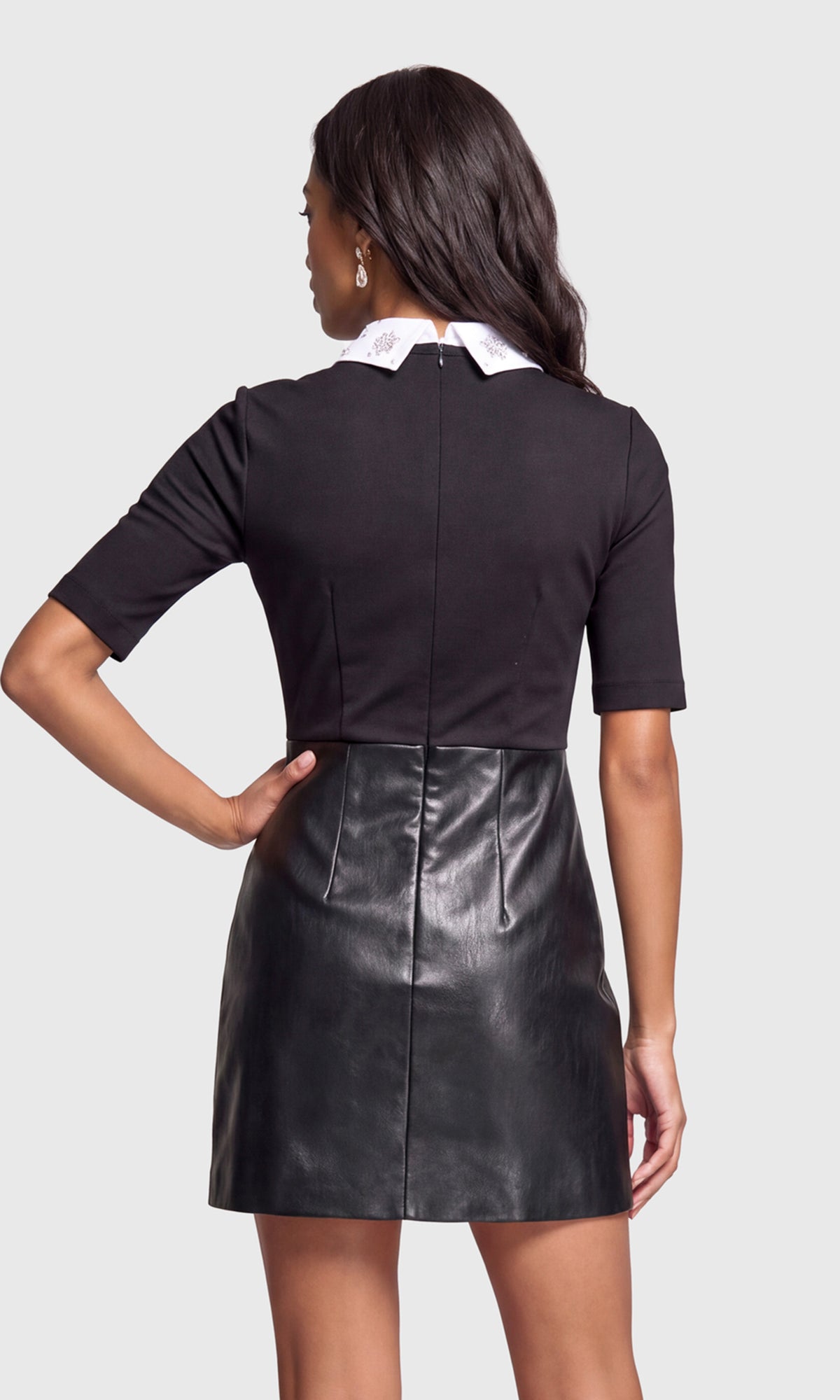 Back view of model wearing a black dress that features a short-sleeve knit top with a white embroidered pointed collar and a black faux-leather mini skirt.