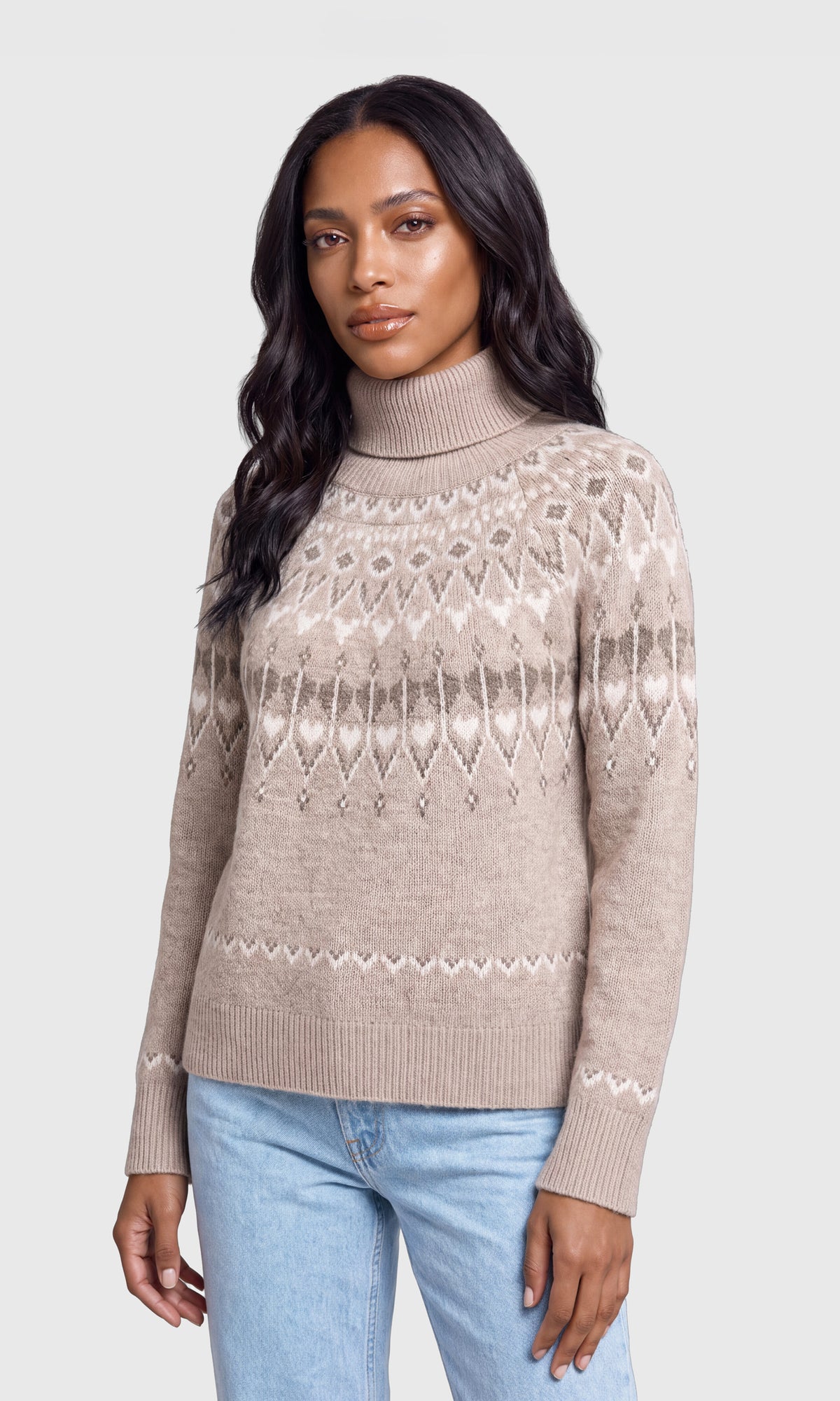 A woman wearing a beige Fair Isle knit turtleneck sweater with light blue jeans, photographed against a solid light gray background.