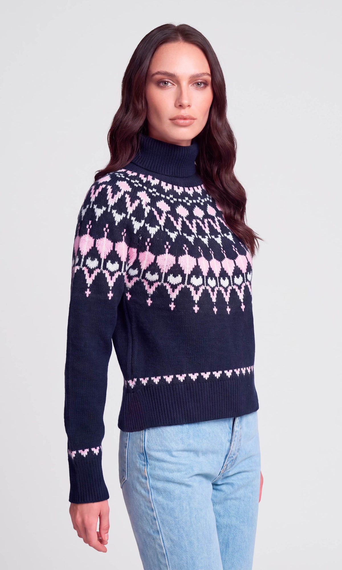 A woman modeling a navy blue turtleneck sweater with a pink and white geometric Fair Isle pattern across the chest and sleeves, paired with light blue jeans.