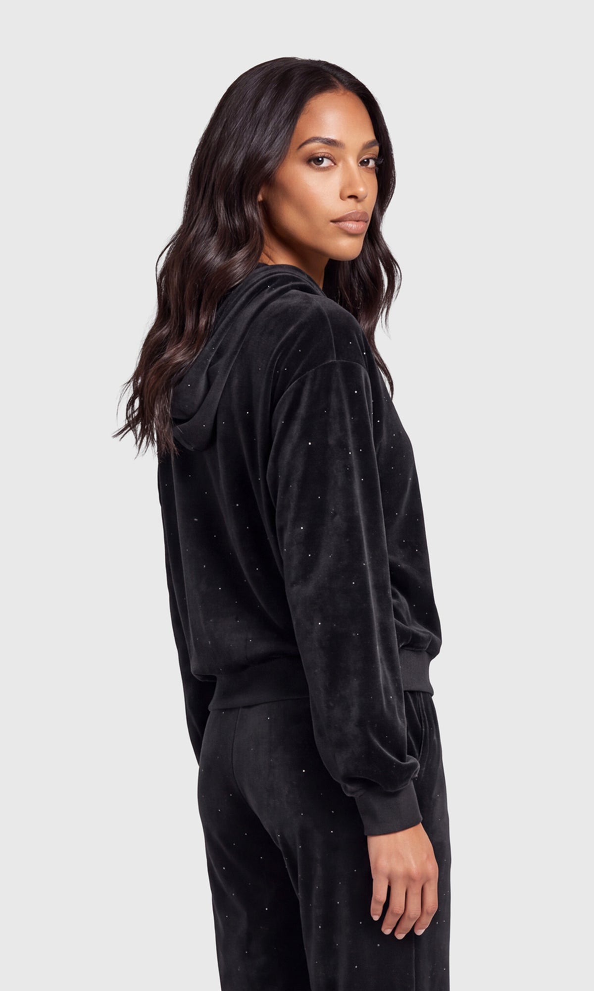 Side view of model wearing a black velvet two-piece loungewear set with subtle silver rhinestone embellishments, consisting of a long-sleeve hoodie and matching pants.