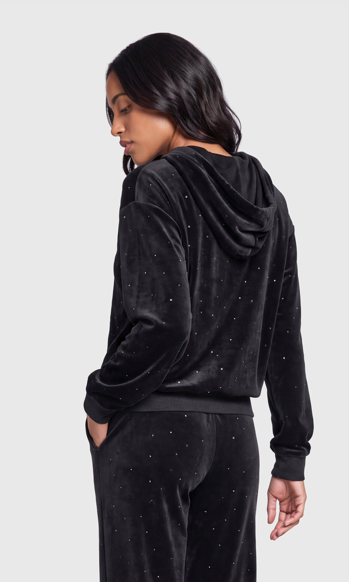 Back view of model wearing a black velvet two-piece loungewear set with subtle silver rhinestone embellishments, consisting of a long-sleeve hoodie and matching pants.
