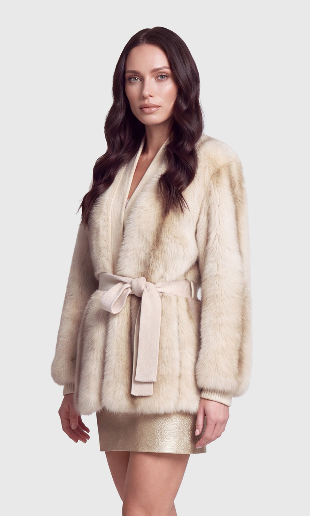 A woman modeling a plush beige fur wrap coat with a matching fabric tie belt, paired with a metallic gold mini skirt.