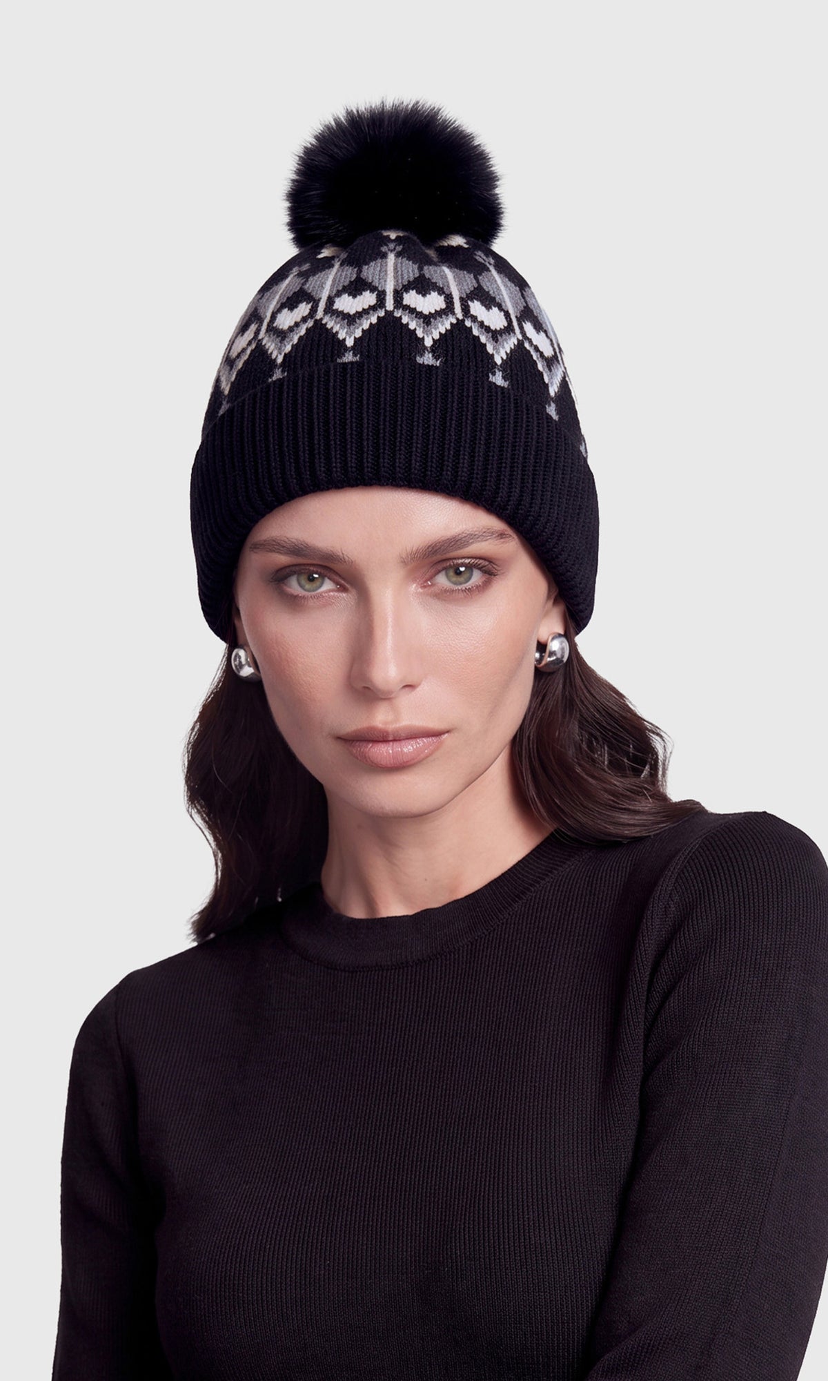 A woman wearing a black knit beanie with a white geometric pattern and a black pom-pom, paired with a black ribbed top.