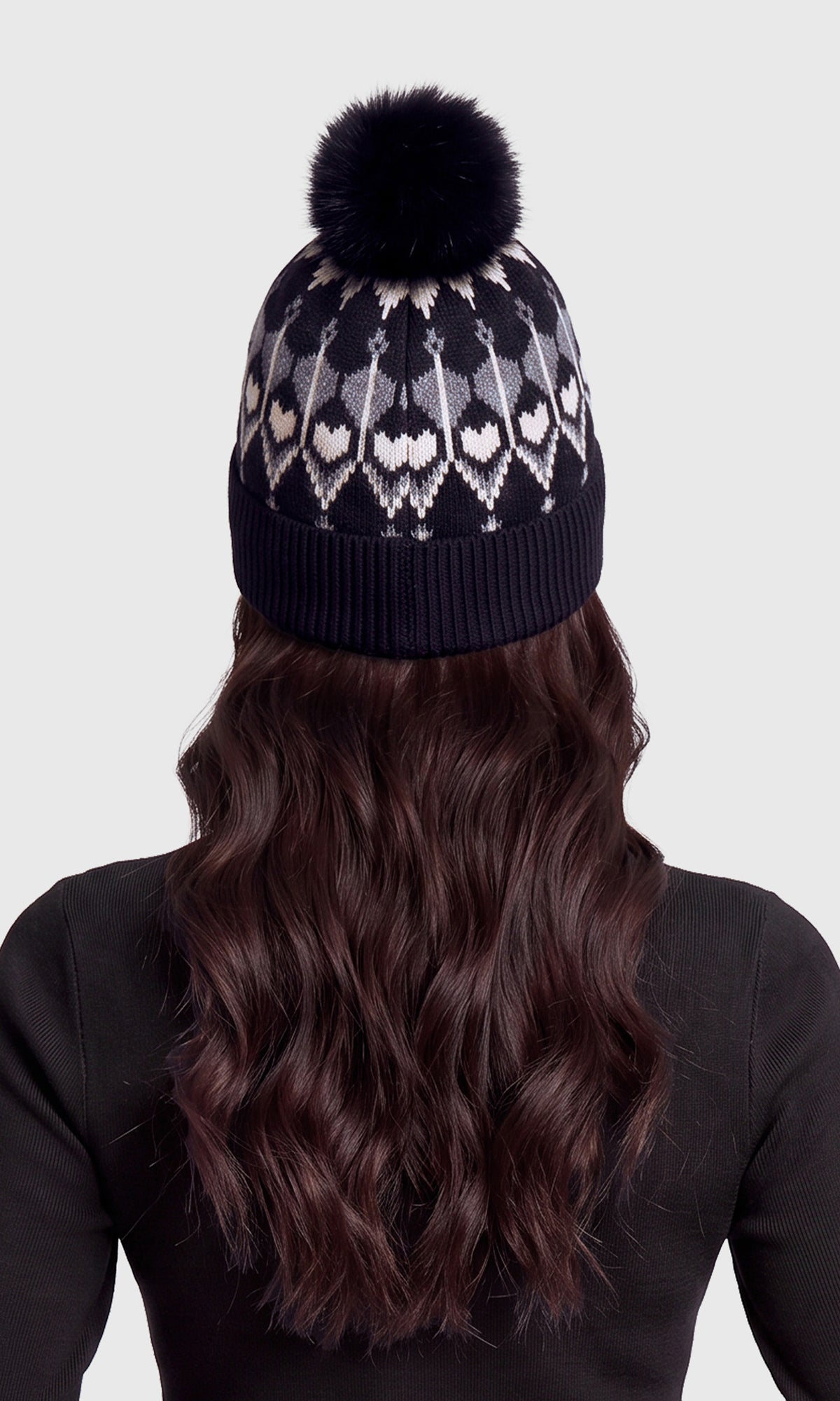 Back view of model wearing a dark navy knit beanie with a black pom-pom and a white and gray geometric heart pattern. 