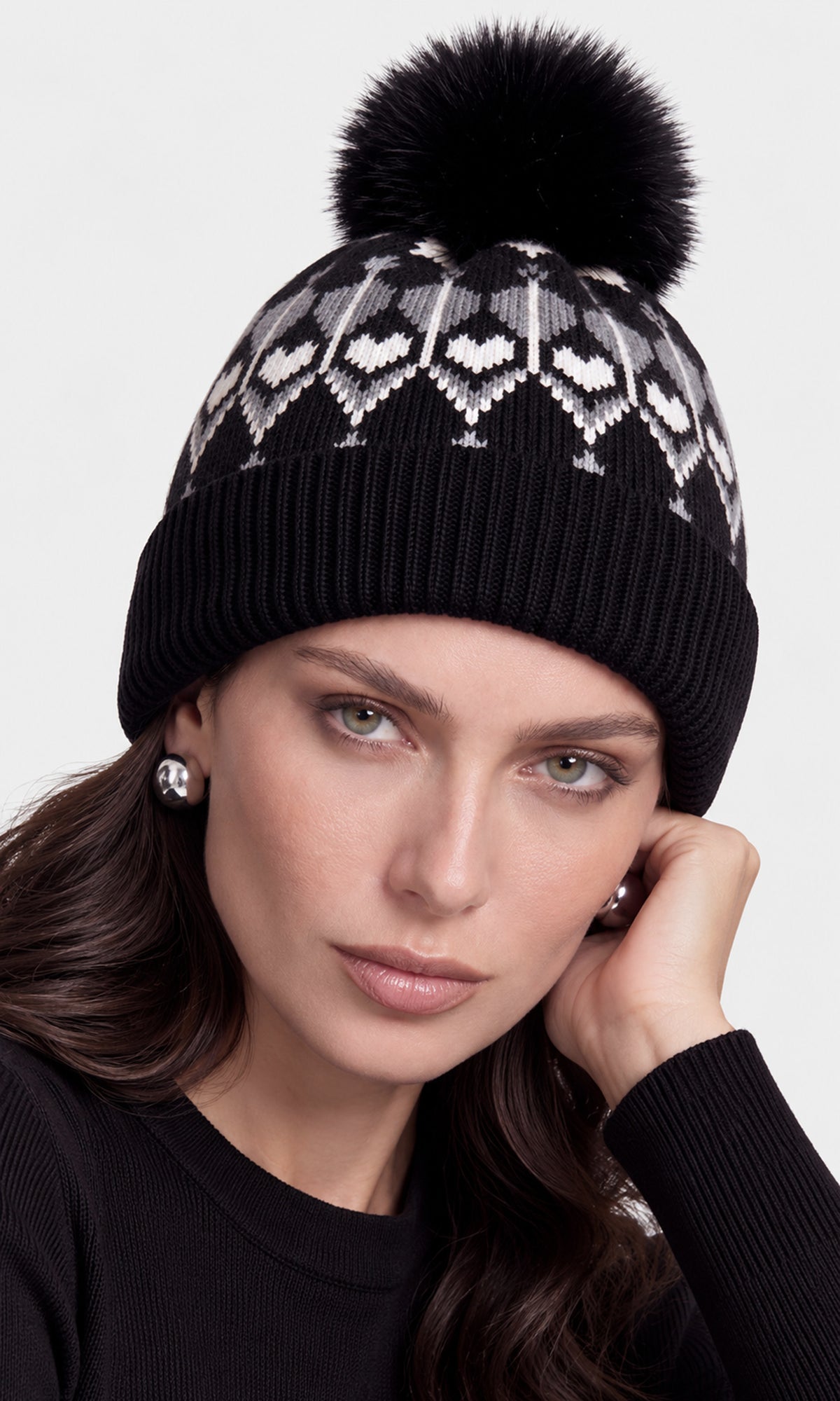 A close-up of a woman wearing a black knit beanie with a white geometric heart pattern and a large black faux-fur pom-pom.