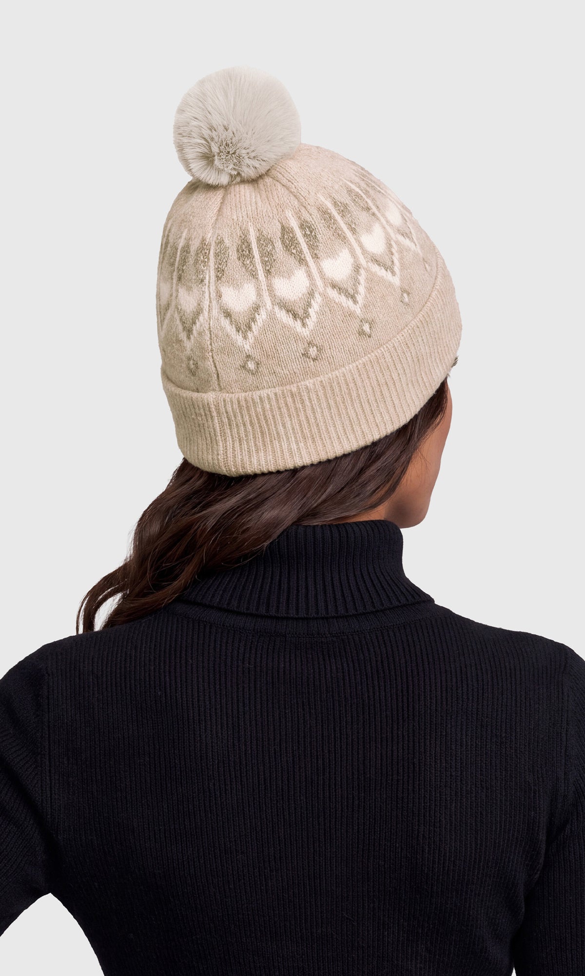 A woman shown from the back wearing a beige knit pom-pom beanie with a subtle heart and diamond pattern, paired with a black ribbed turtleneck sweater.