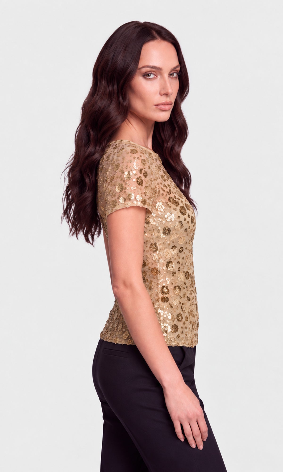 A woman with long dark wavy hair is shown in side profile wearing a short-sleeve gold sequined top and black pants against a light gray studio background.