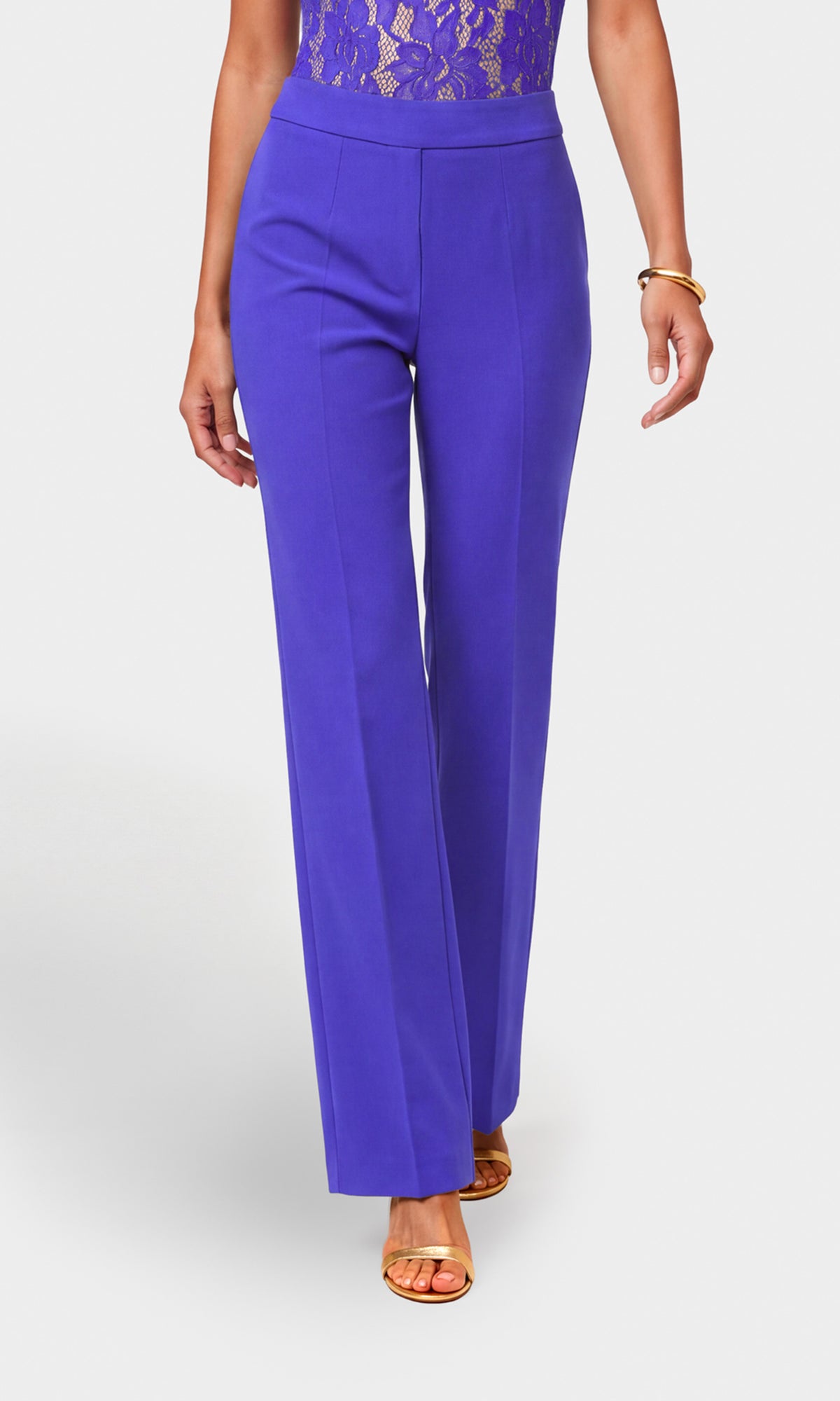 A model wearing high-waisted, wide-leg royal purple trousers paired with a purple lace bodysuit top and gold heeled sandals.